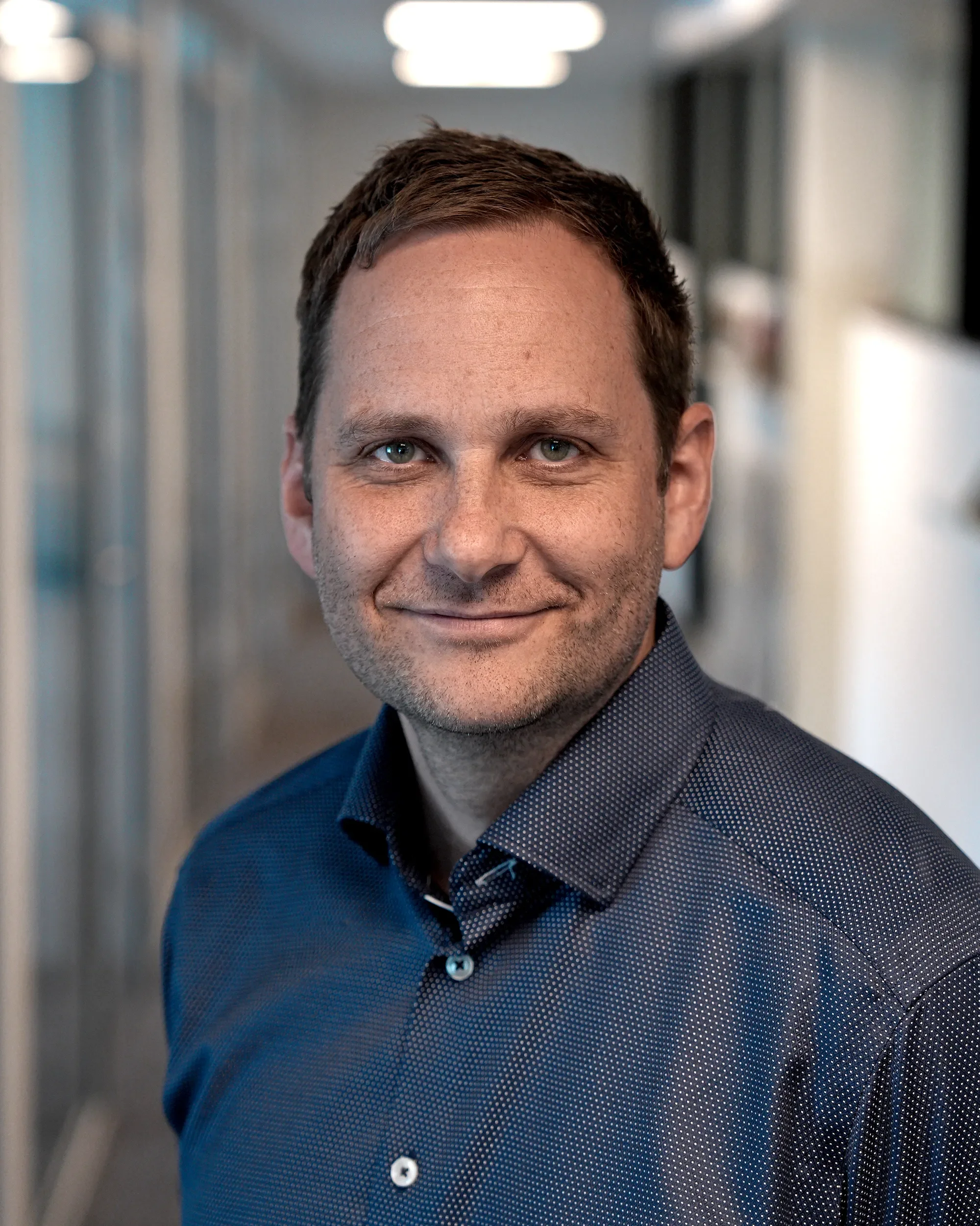 Nærbilde av en smilende mann i dress, med kortbrunt hår, i en kontorbygning.