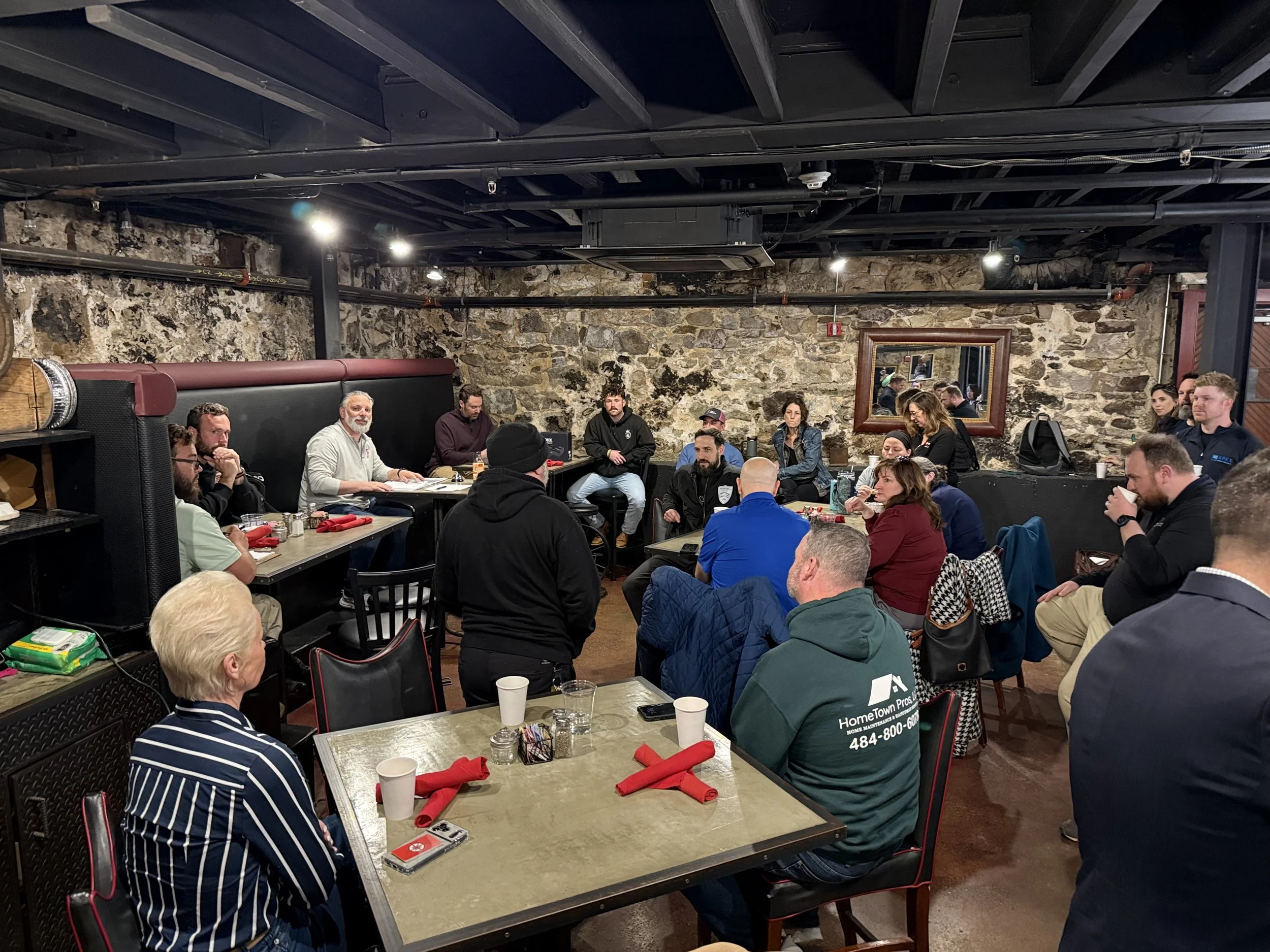 A group of people seated around tables in a rustic restaurant with exposed stone walls, listening to a speaker at the front. The tables have cups, napkins, and small items. There is a large mirror on the back wall and some lighting fixtures on the ceiling.