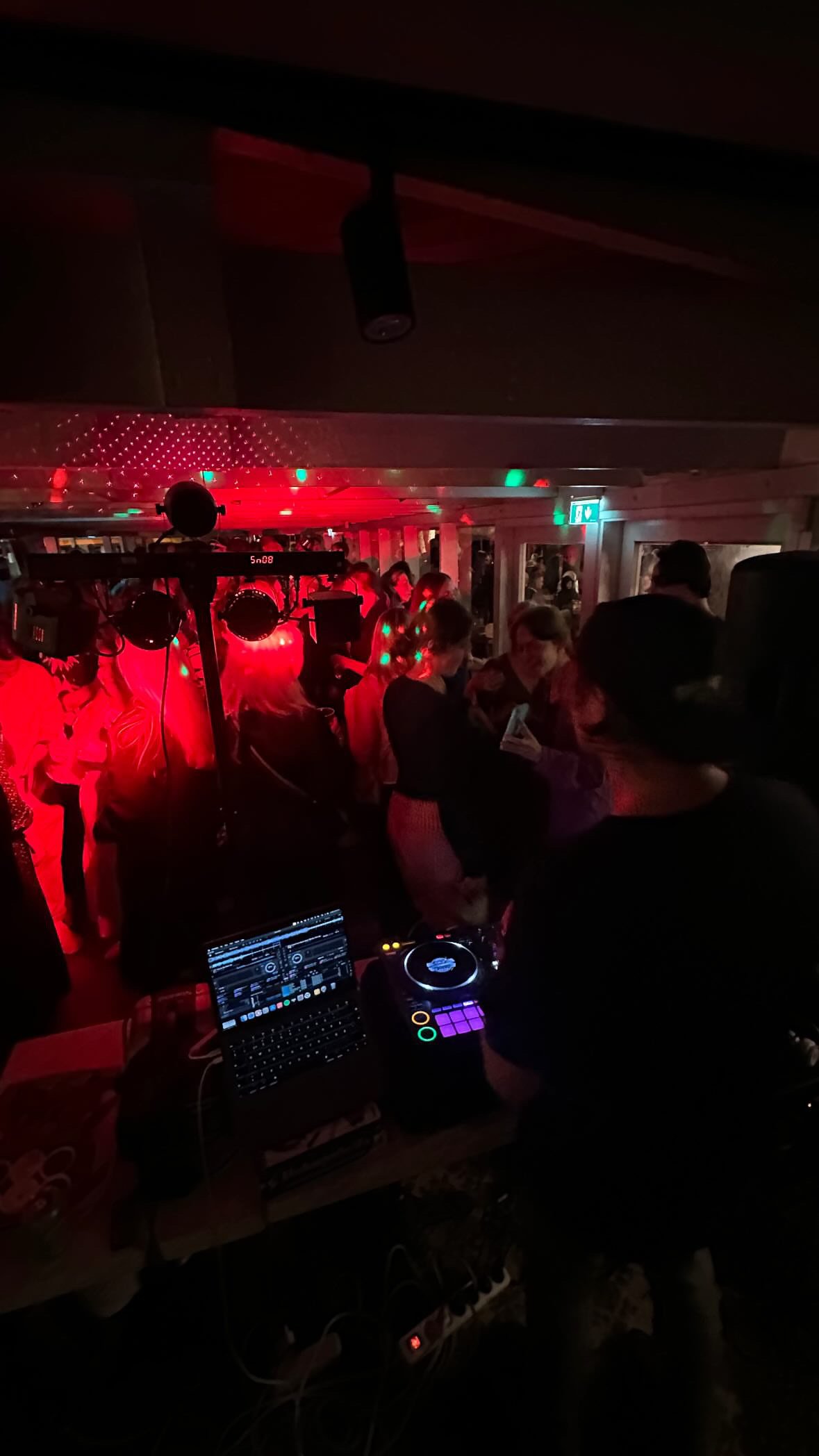 Tanssilattialla on useita ihmisiä, joissa on päällä juhla-asut ja joissa on vilkkuvia värivaloja. DJ-pöydällä on tietokone ja musiikkilaitteita.