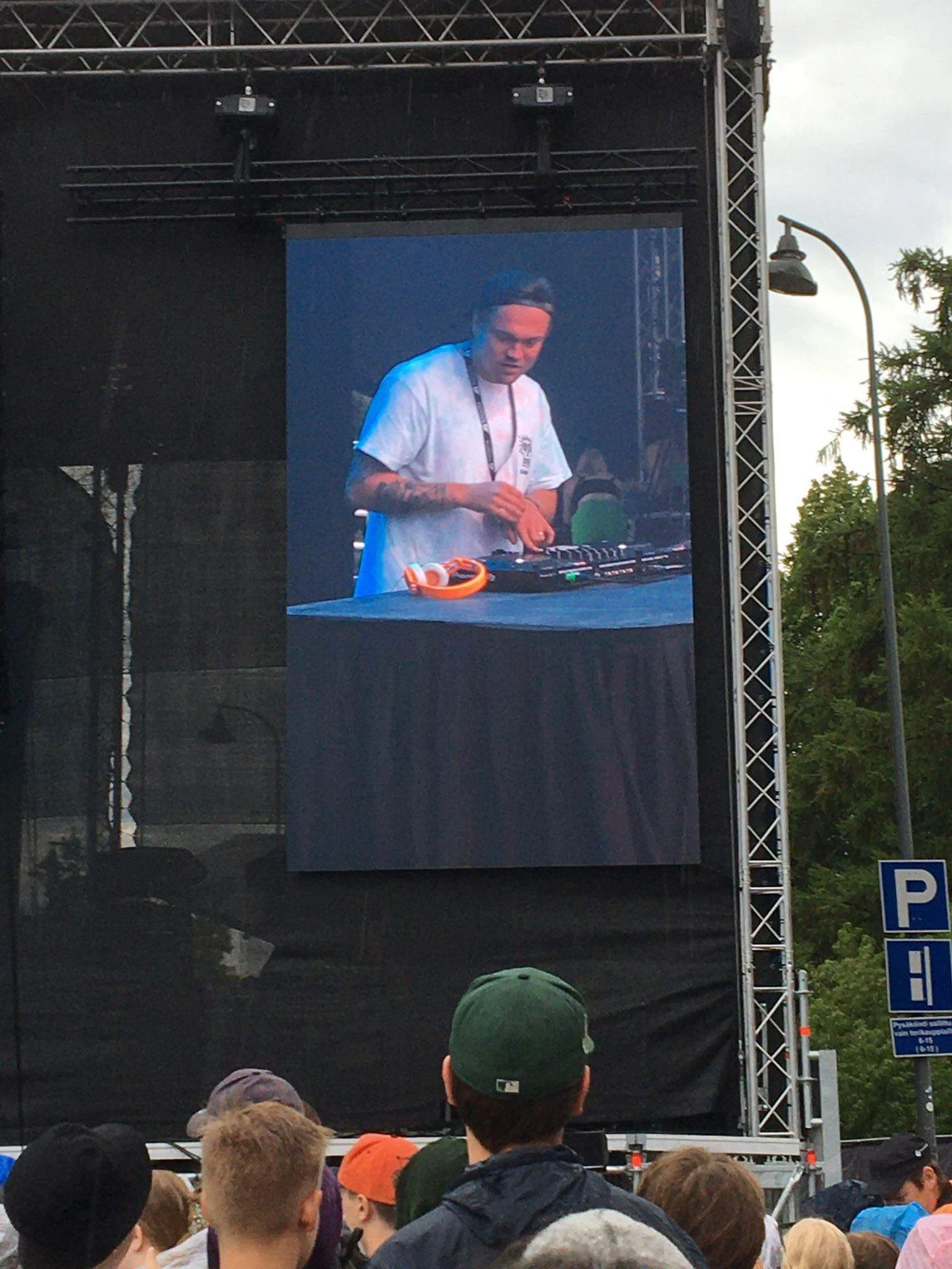 Yleisö katsoo suurta ulkoista videota, jossa DJ soittaa musiikkia. Taustalla näkyvyys puita ja pilvinen taivas.