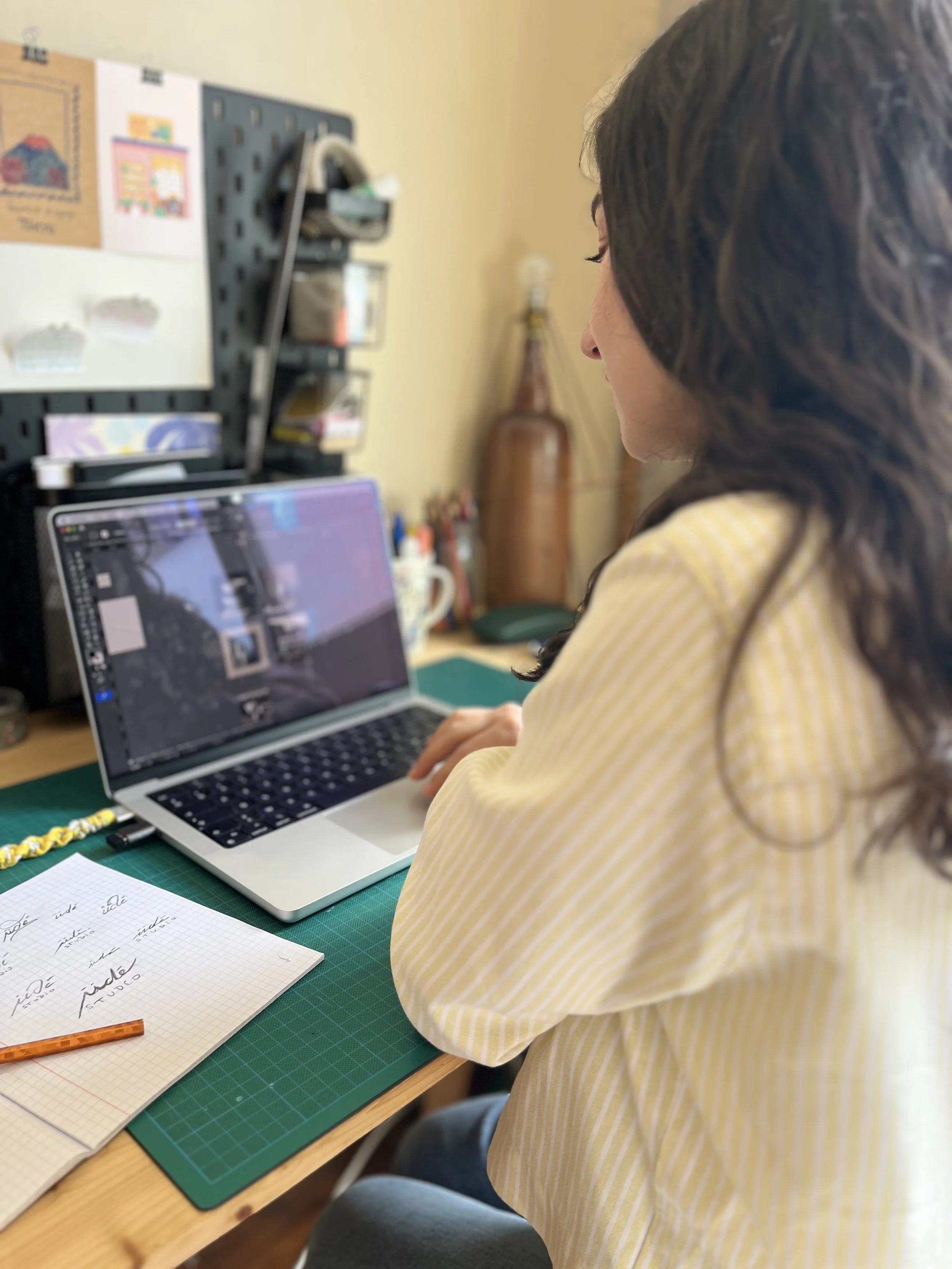 Une femme avec des cheveux bruns foncés, vue de profil, travaille sur un ordinateur portable dans un bureau. Sur la table, il y a un carnet avec des notes manuscrites, un crayon orange, un masque et une règle.