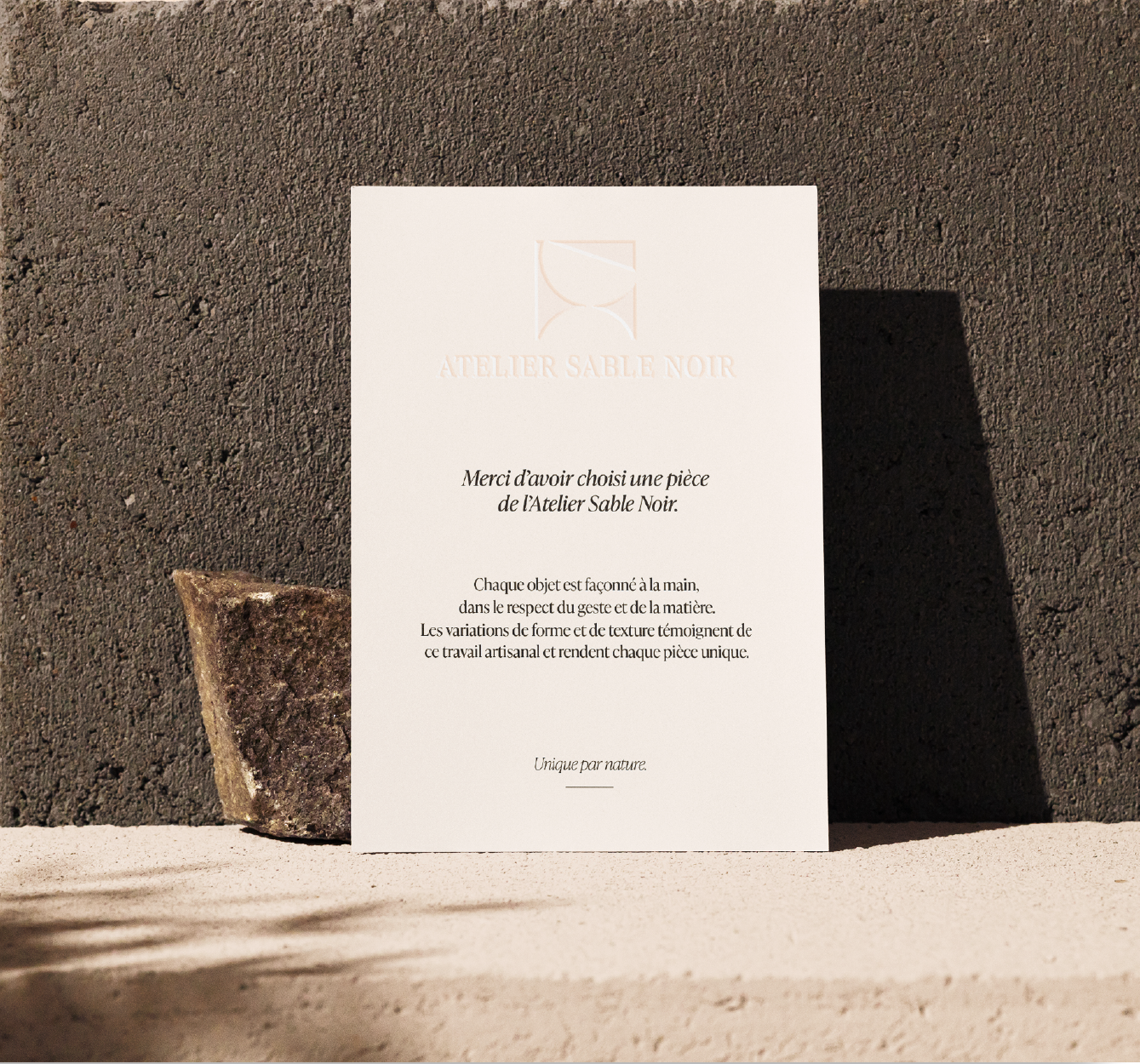 Une carte blanche avec texte en français, posée contre un mur en béton, avec une pierre à gauche.