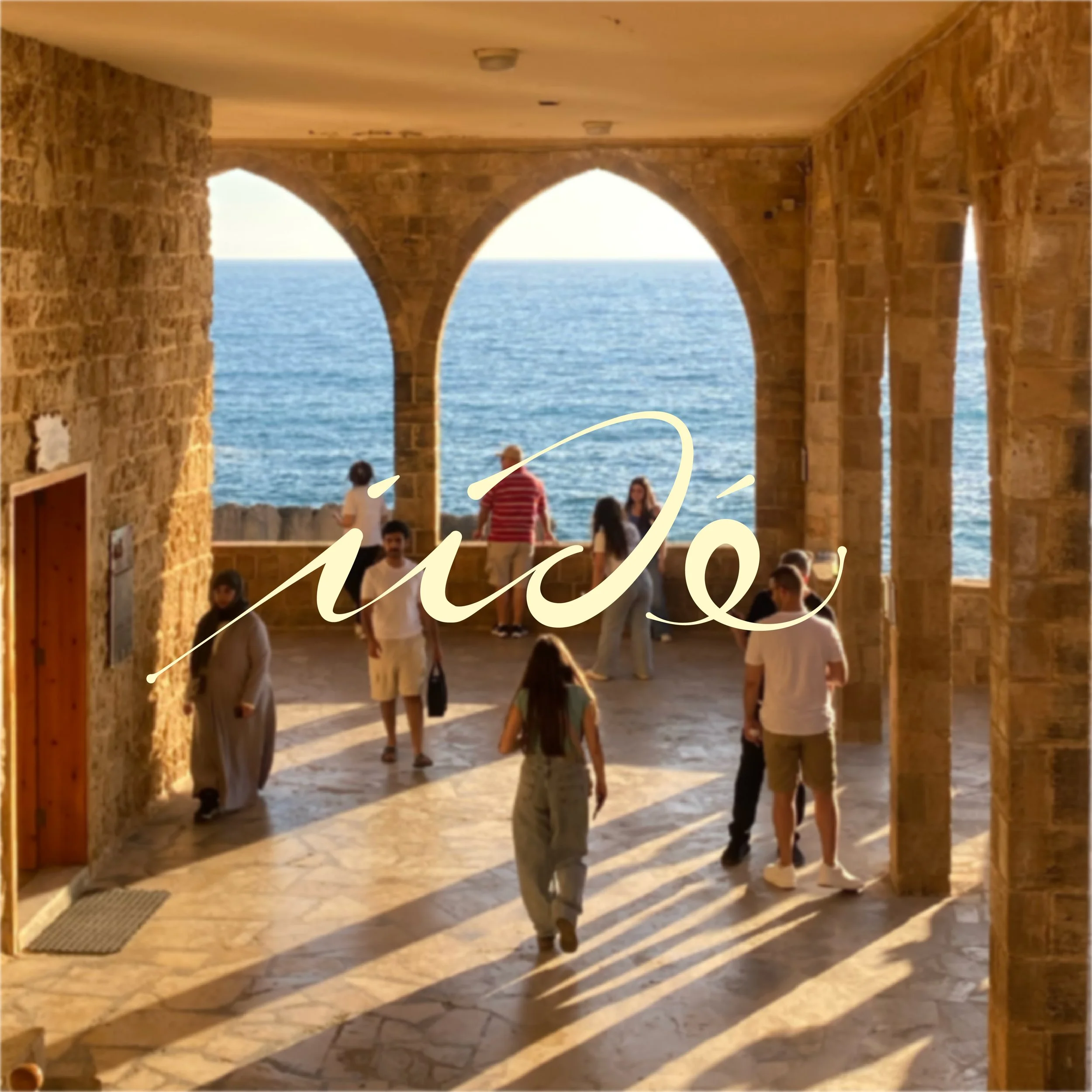 Une vue d'une terrasse avec des arches en pierre donnant sur la mer, avec des personnes debout et assises dans l'ombre, profitant du coucher du soleil.