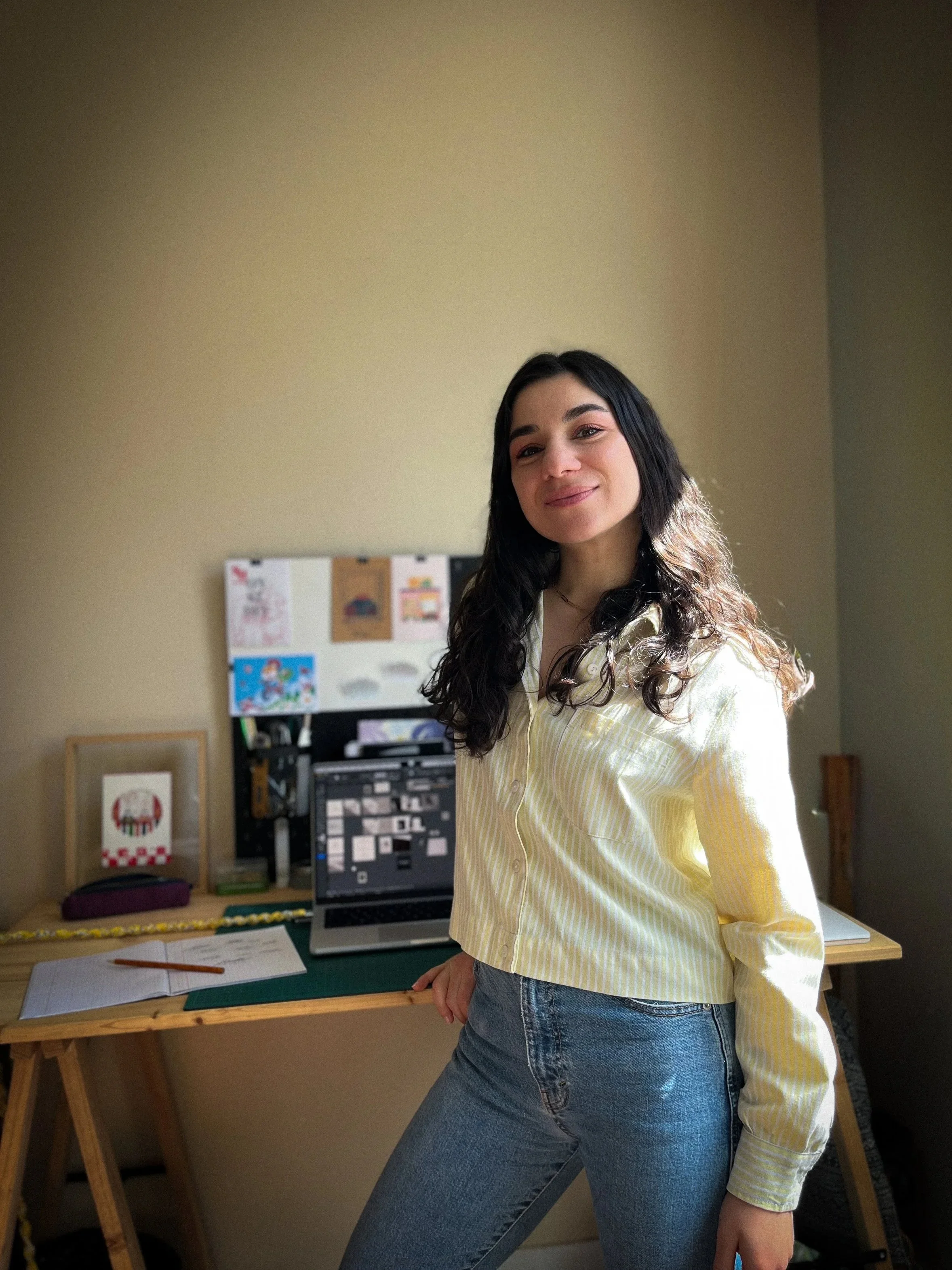 Une jeune femme aux longs cheveux ondulés et à la peau claire pose dans un bureau, portant une chemise jaune à rayures et un jean.