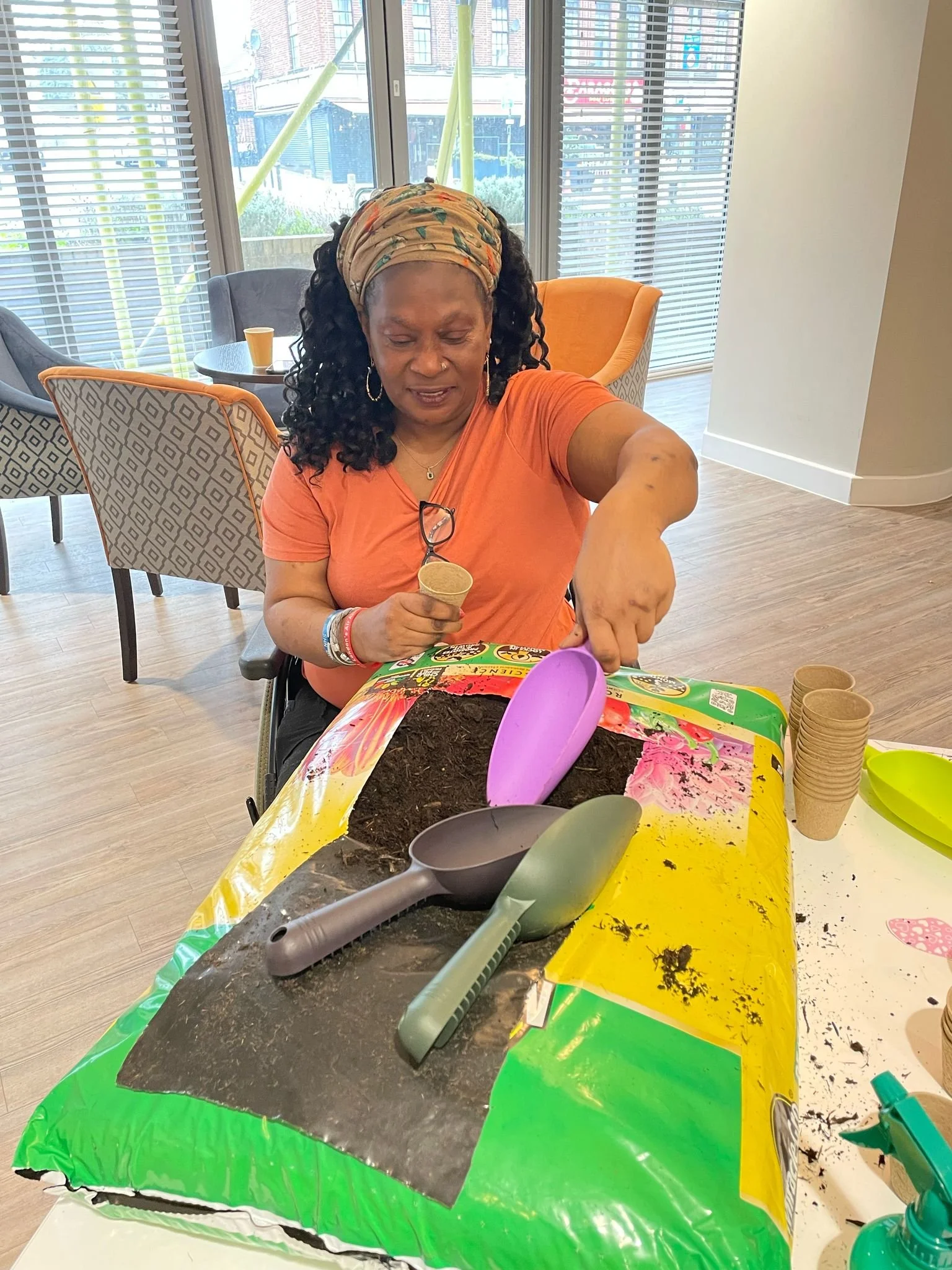 A woman sitting at a table, grabbing soil from a bag, with gardening tools and small plant pots on the table in a bright room with large windows.