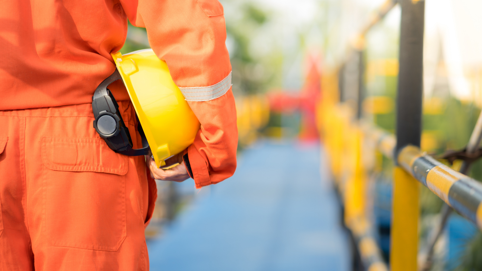 Close-up of a construction worker wearing orange safety uniform, holding a yellow safety helmet, standing on a construction site near yellow and black safety barriers.
