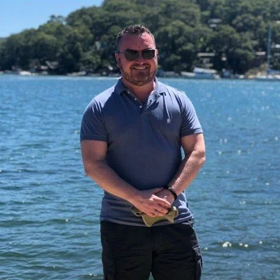 A man with sunglasses smiling and standing by a body of water with trees in the background.
