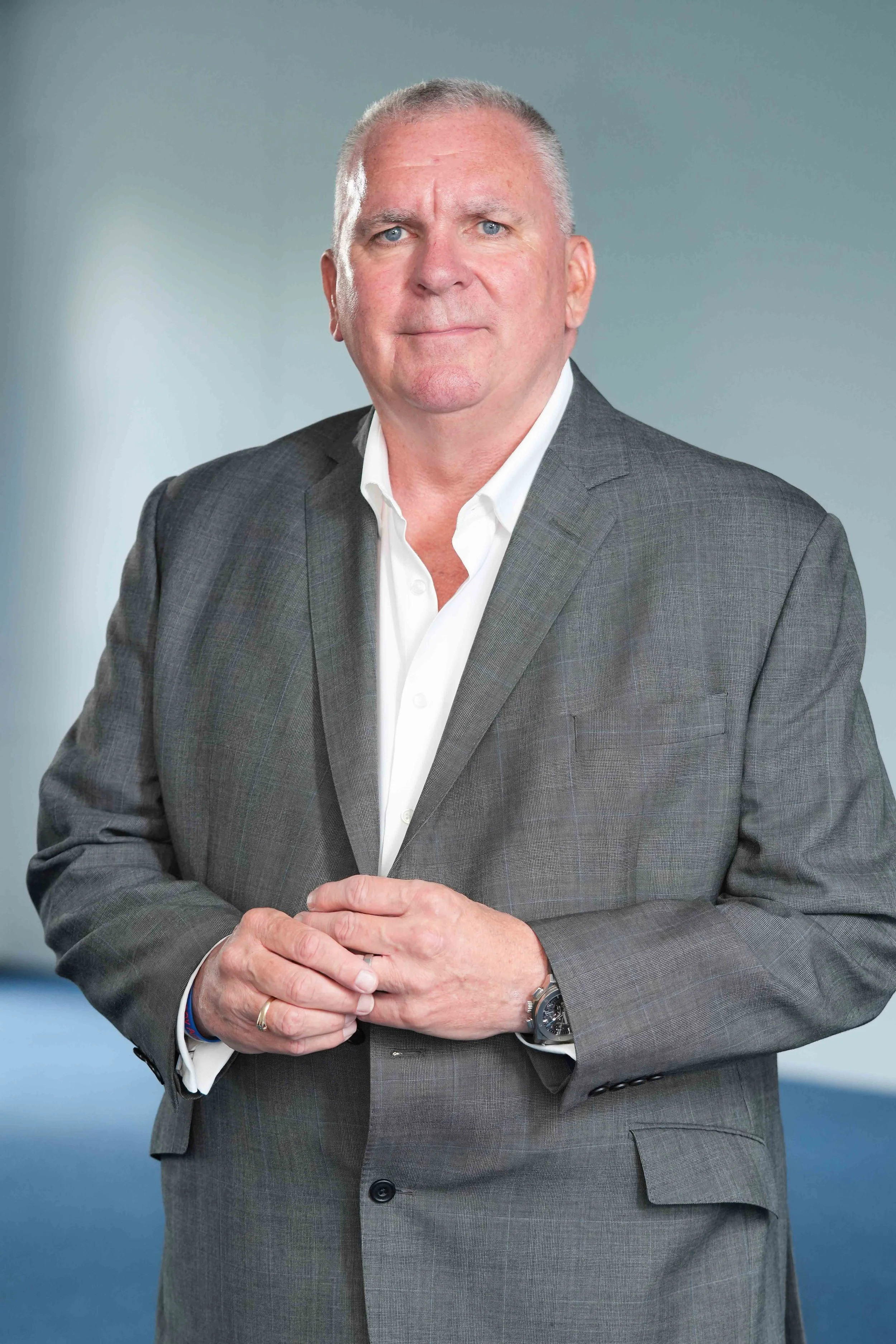 A man in a grey suit and white shirt, standing with his hands clasped in front of him, against a plain background.