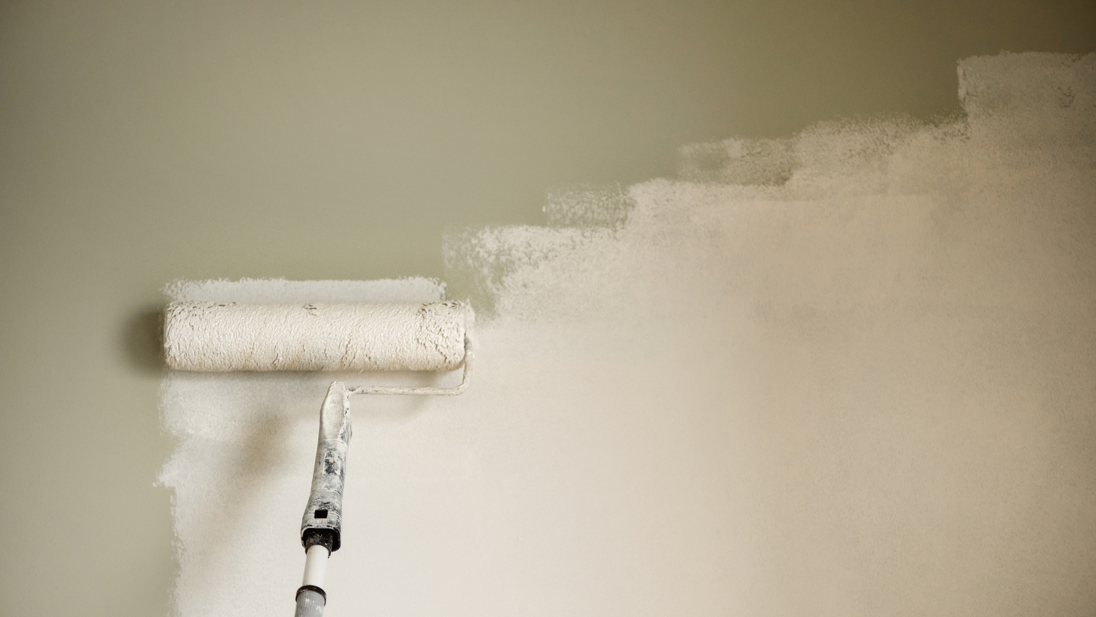 A person using a paint roller to paint a wall a light beige color.