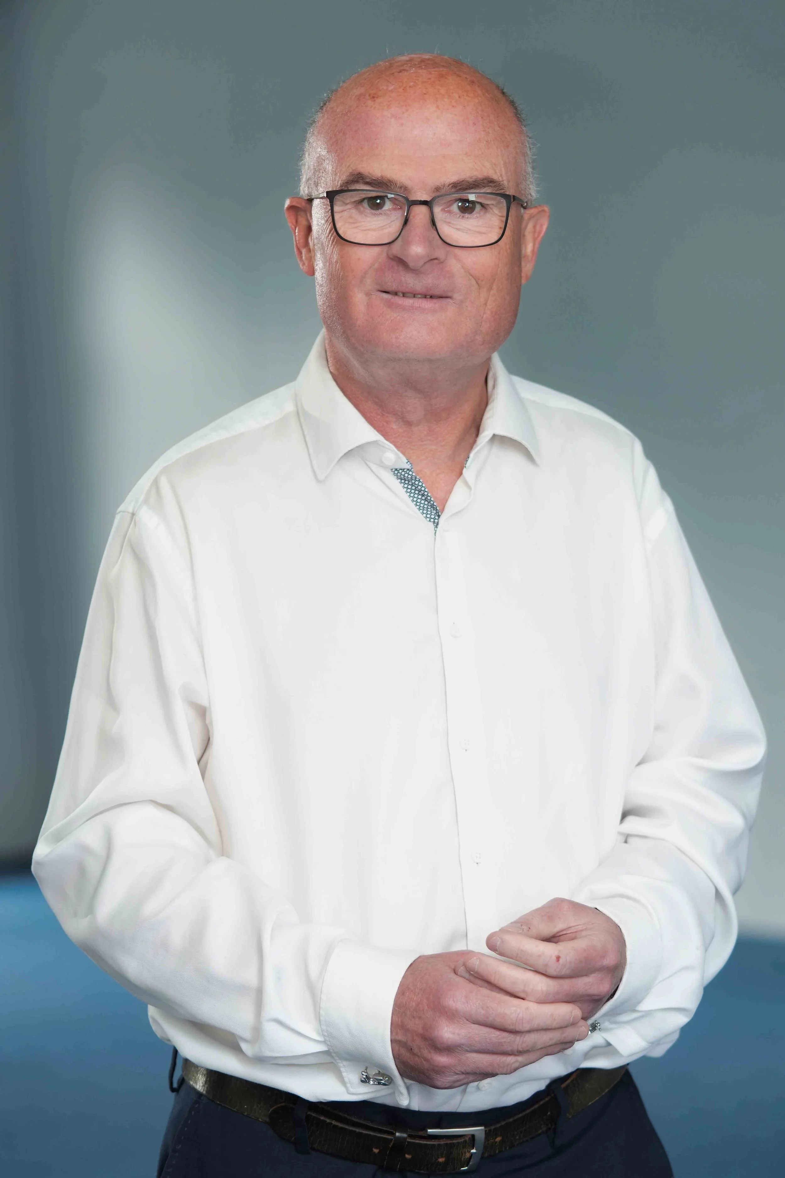 A man wearing glasses and a white dress shirt, posing against a neutral background.