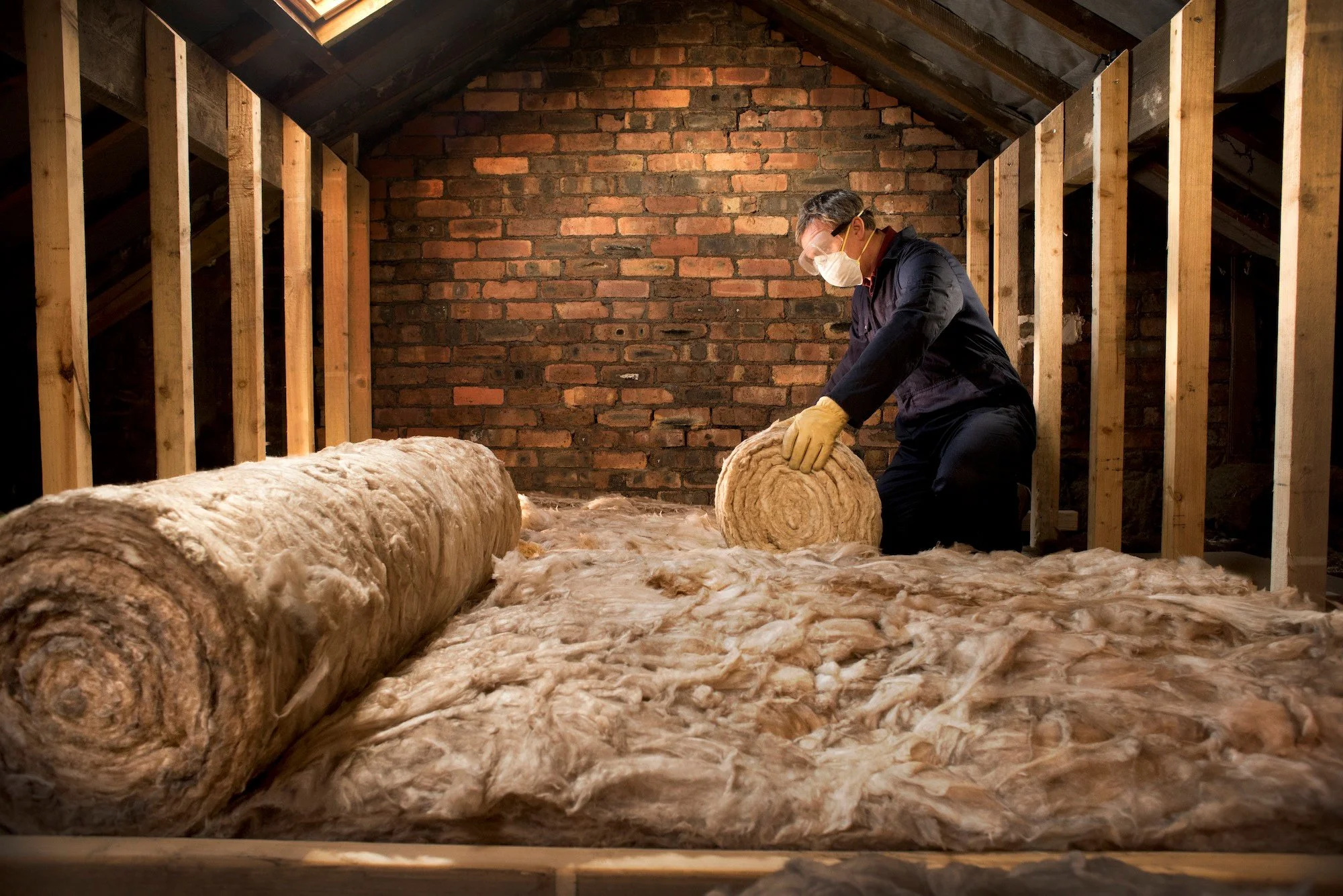 A person wearing a mask and gloves working with insulation material in an unfinished attic space with wood framing and red brick wall.