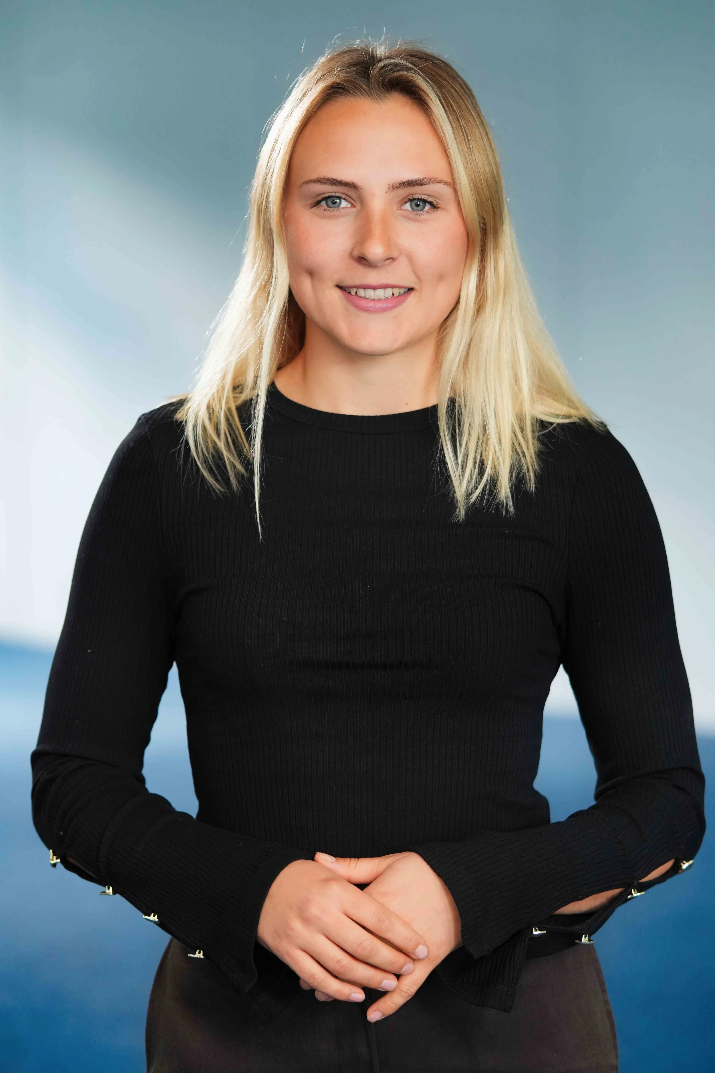 A woman wearing a black long-sleeve top, smiling gently, holding her hands together in front of her against a gradient blue background.