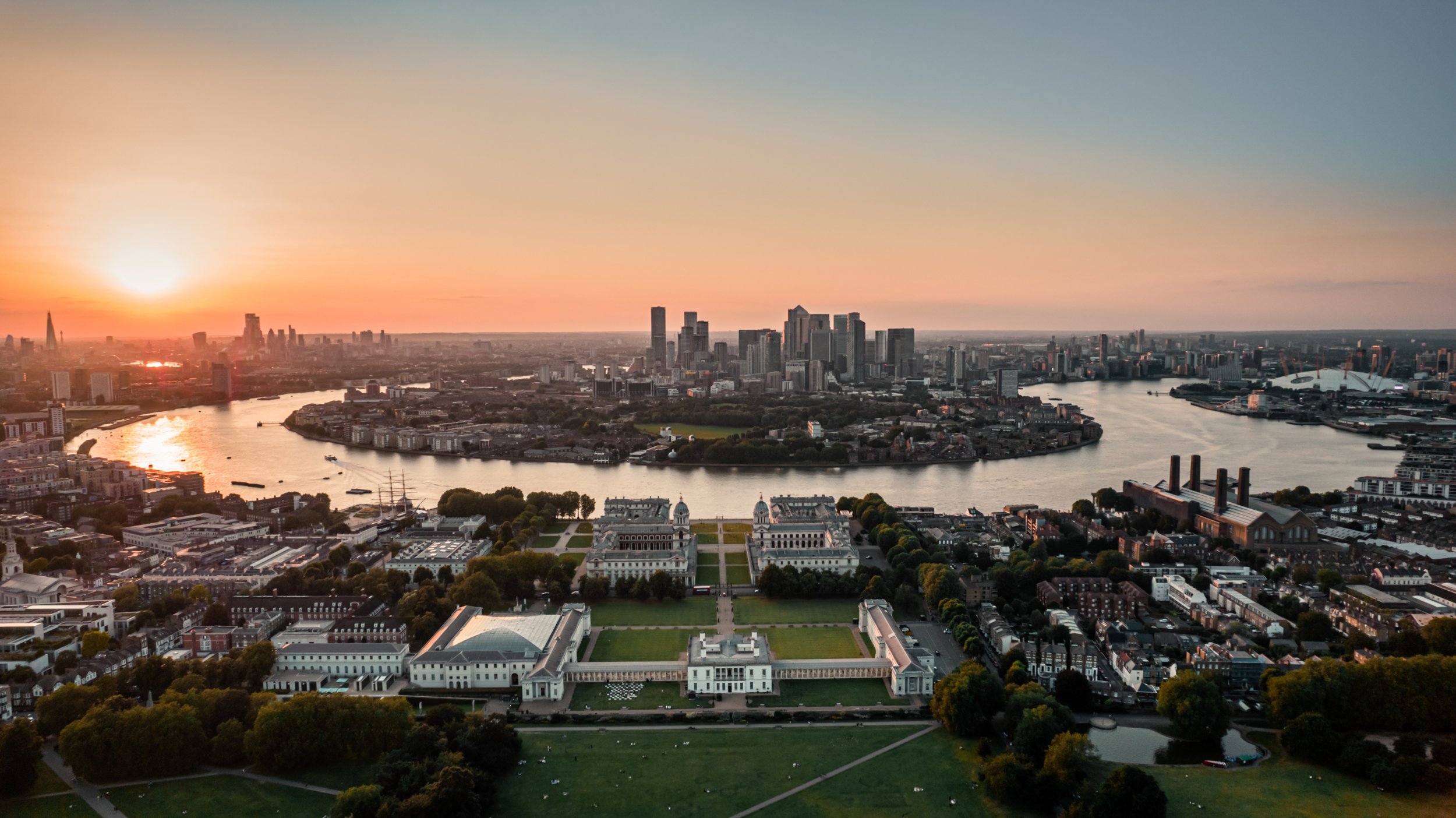 An aerial view of London at sunset, featuring the River Thames flowing through the city, with modern skyscrapers in the background and historic buildings in the foreground, under a pastel-colored sky.