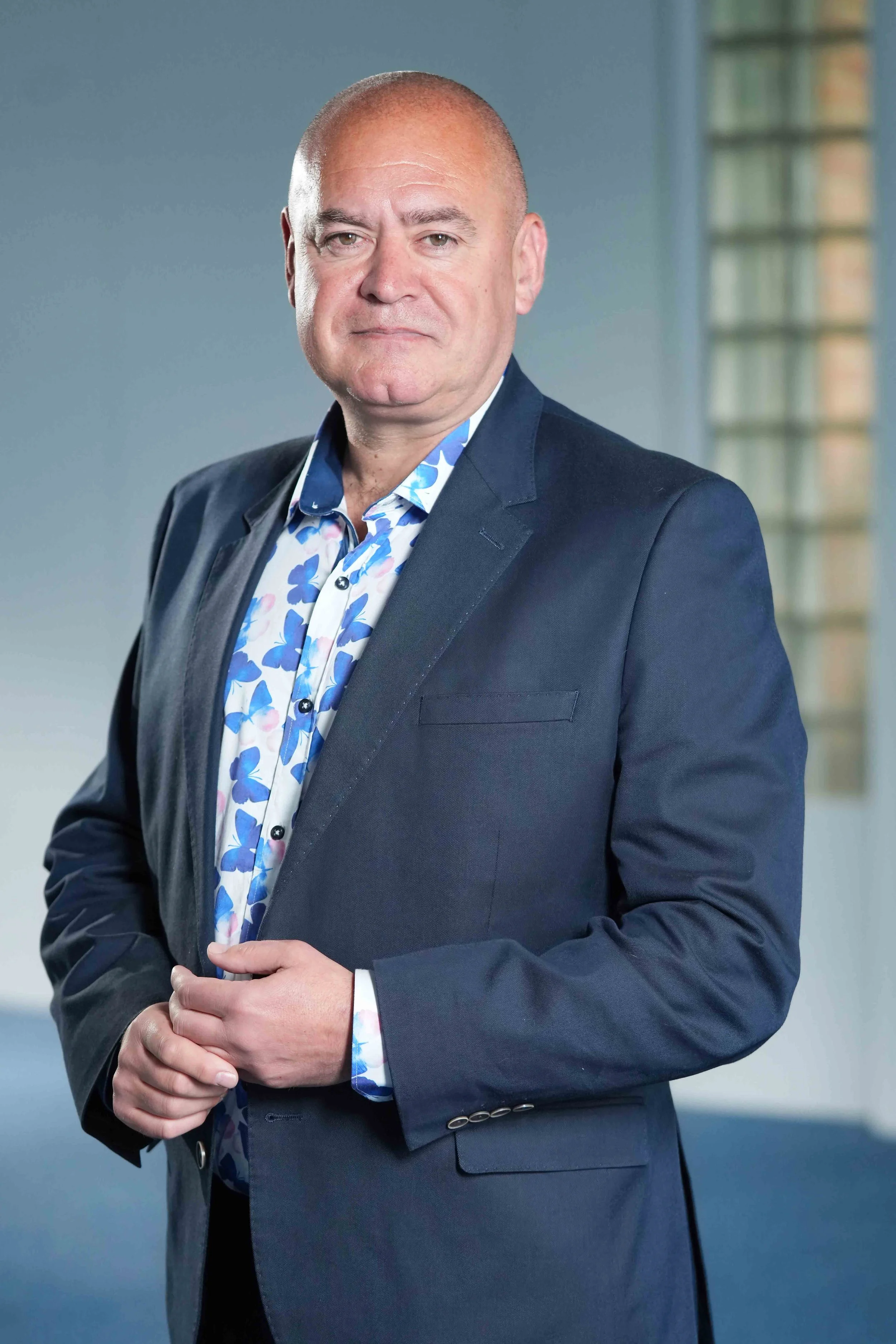 A  man in a blue suit and a floral shirt, standing with his hands clasped in front of him, looking directly at the camera, in an indoor setting.