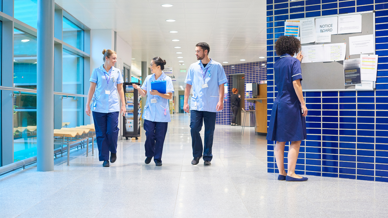 Four healthcare professionals, three nurses and one staff member, walking and conversing in a hospital corridor near a notice board.