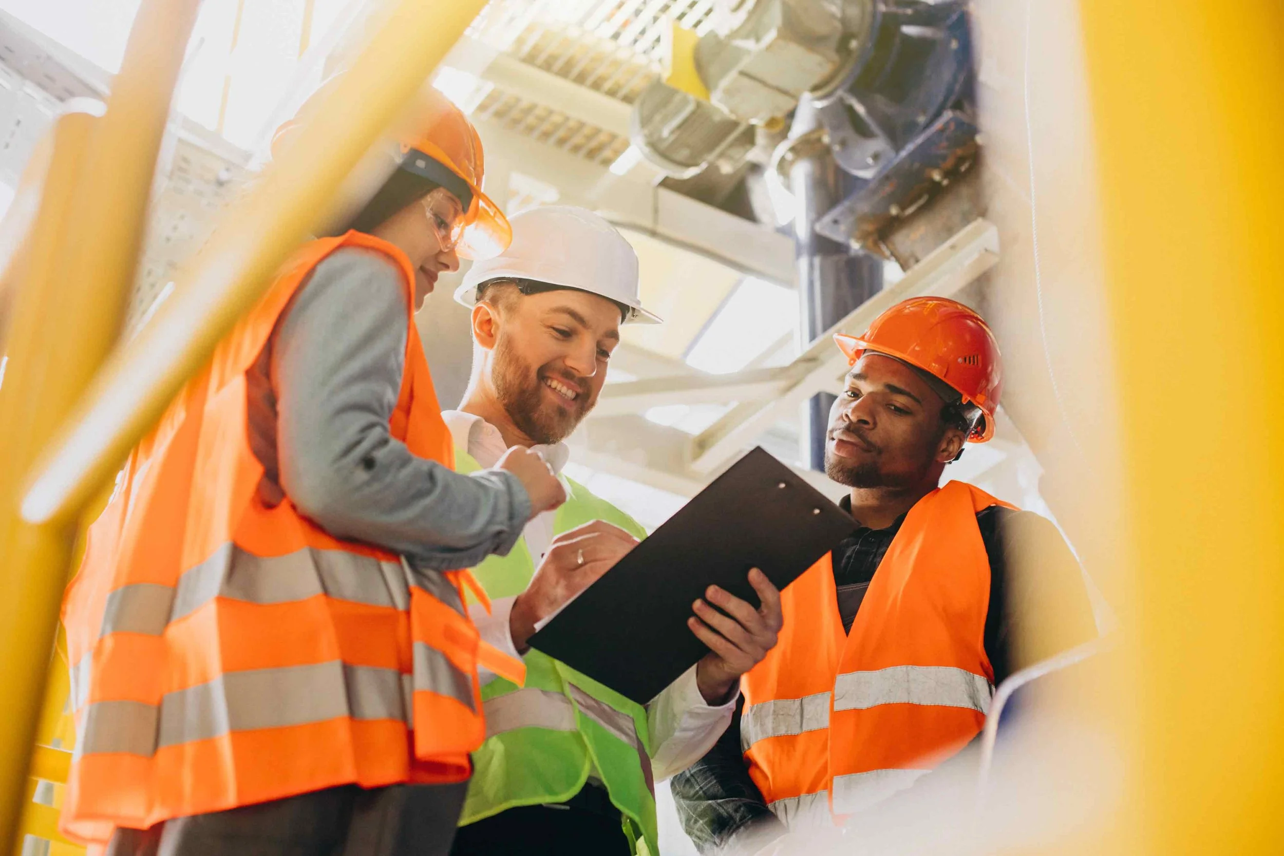 Three construction workers wearing orange and yellow safety vests and hard hats look at a clipboard inside a building.