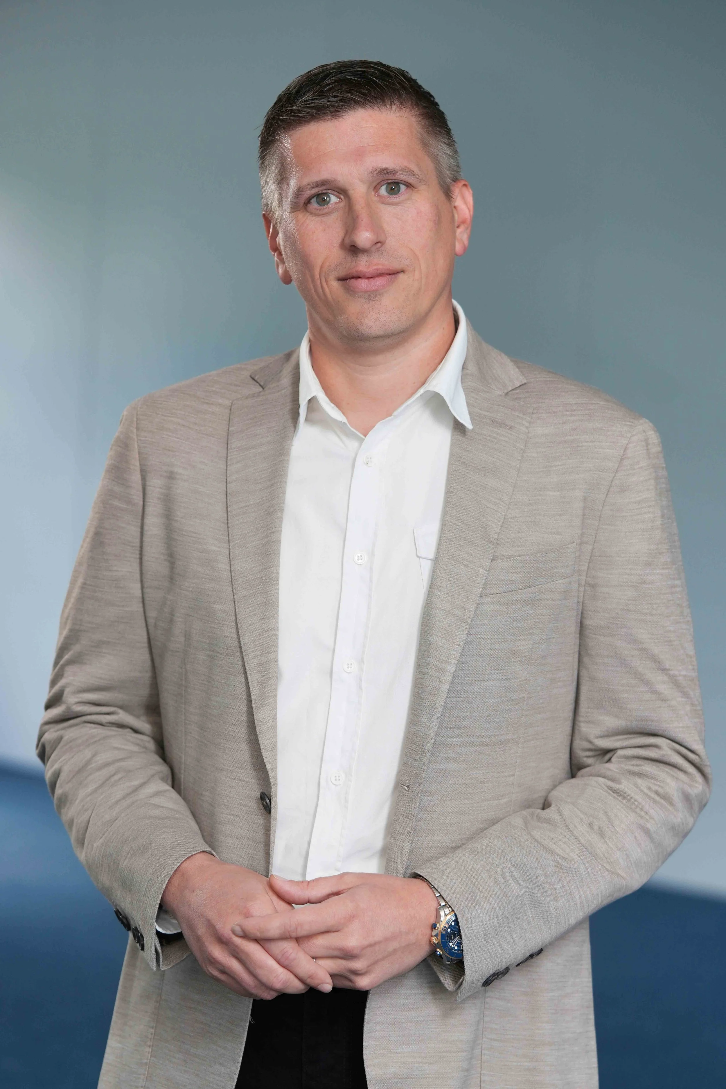A man in a beige blazer and white shirt standing with his hands clasped in front of him against a blue background.