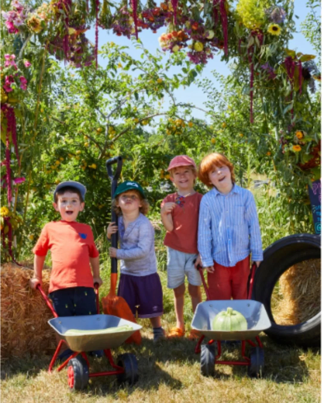 Four children standing in a garden under a floral arch, holding small wheelbarrows with pumpkins inside, surrounded by hay bales and a tire, on a sunny day.