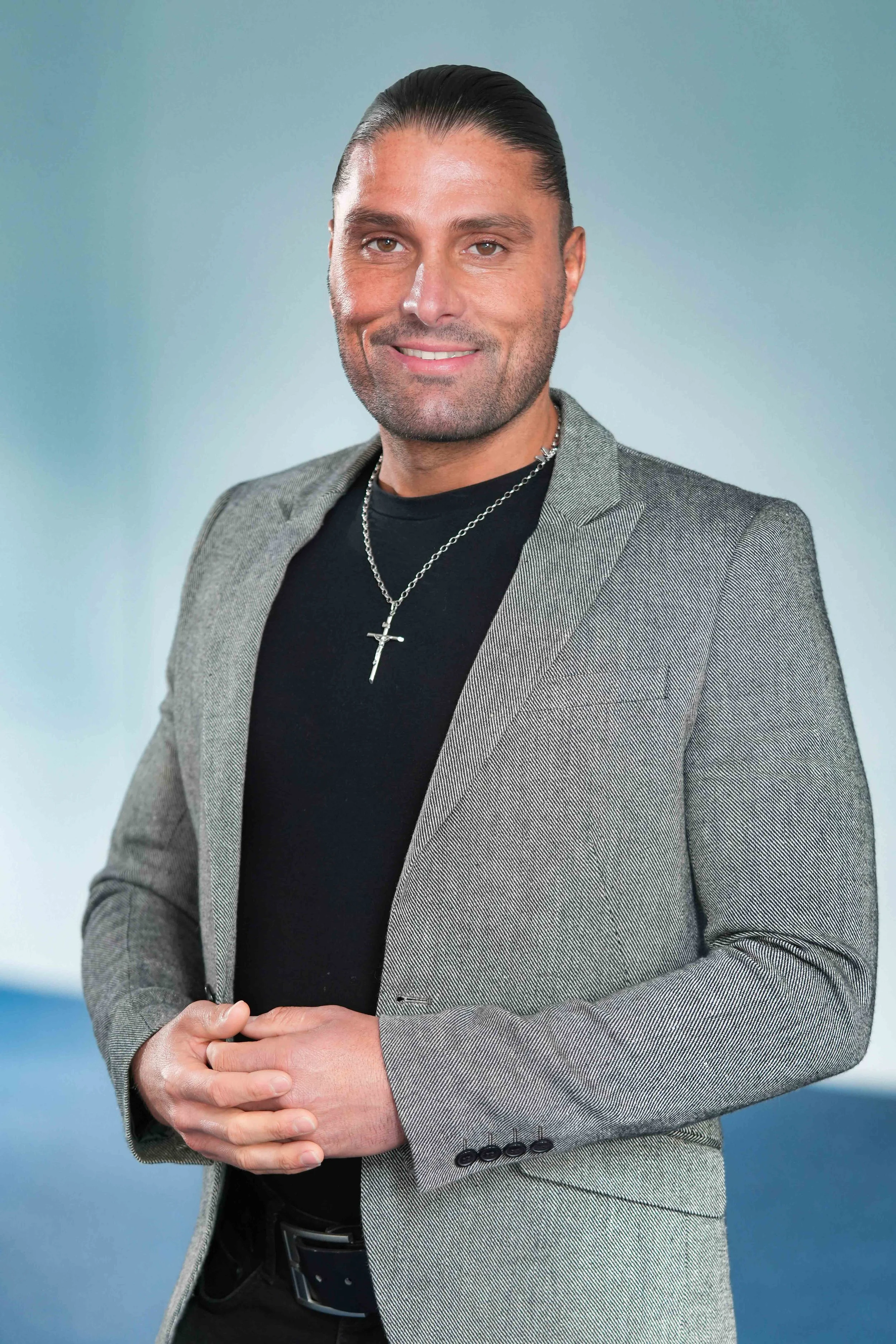 A man with slicked-back dark hair, wearing a grey blazer over a black shirt with a cross necklace, smiling against a blue and green gradient background.
