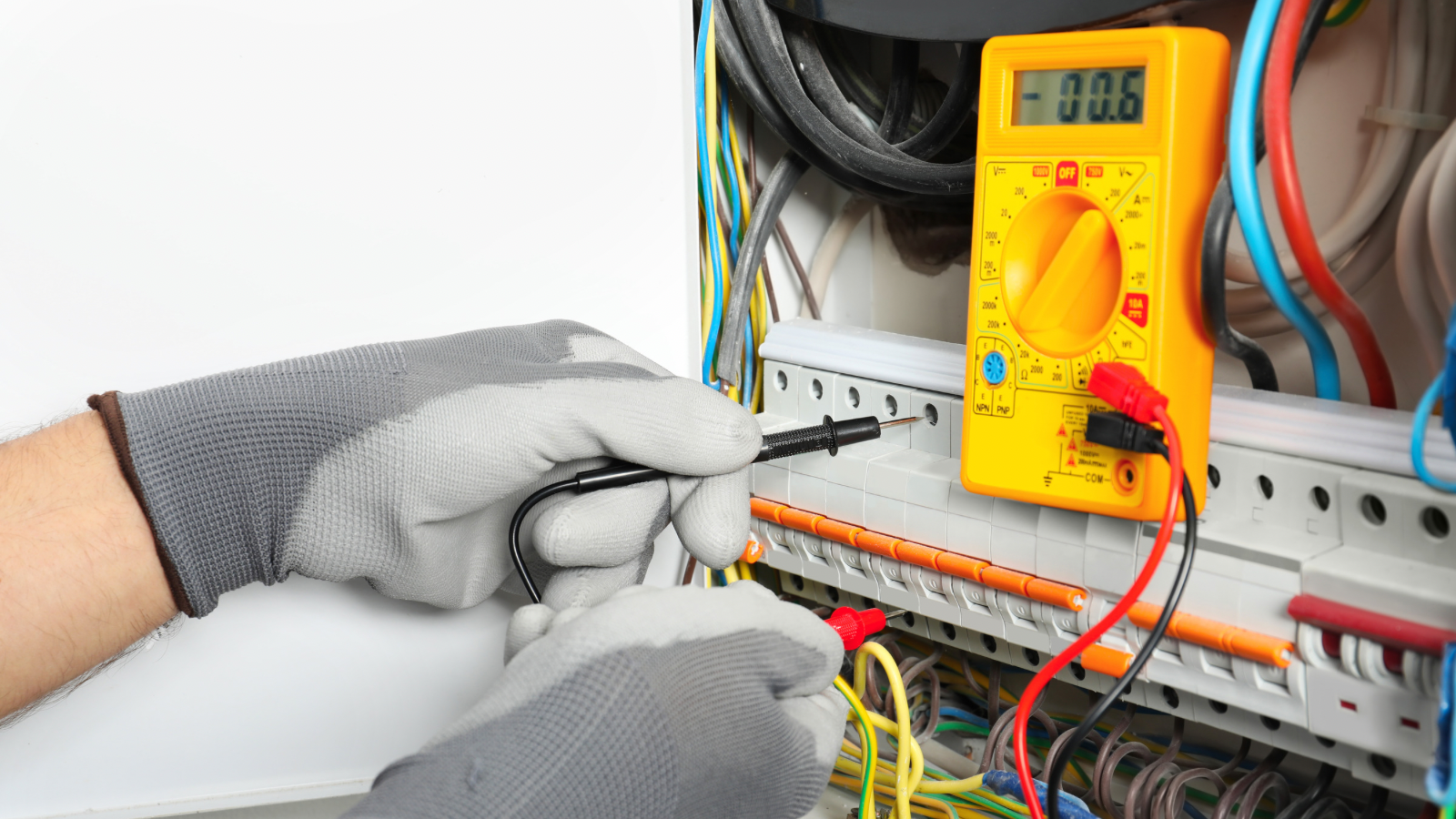 An electrician wearing grey gloves using a screwdriver to test or repair wiring inside an electrical panel, with a yellow multimeter placed on the panel.