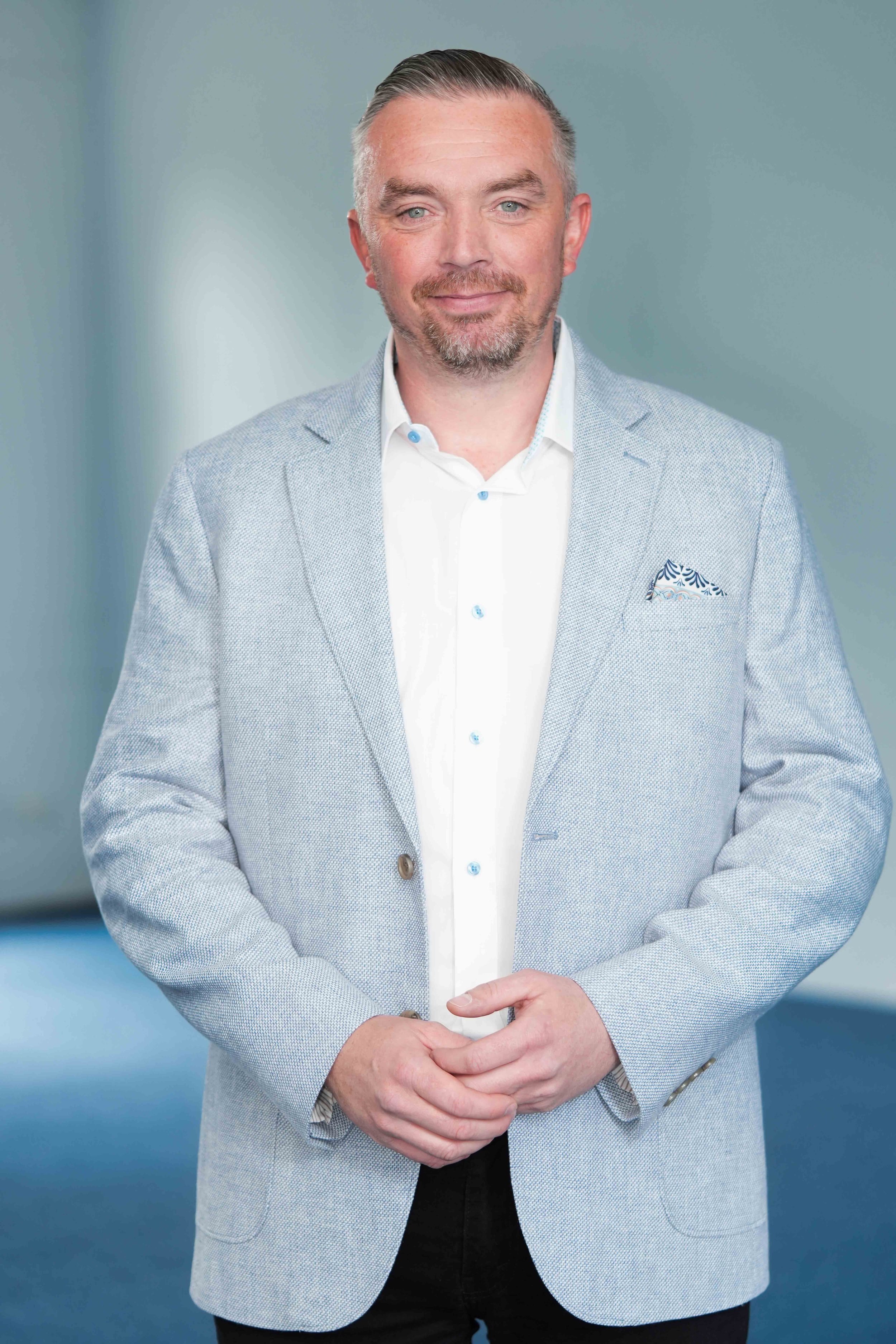 A man with wearing a light grey blazer over a white dress shirt, standing in front of a blurred blue-grey background.