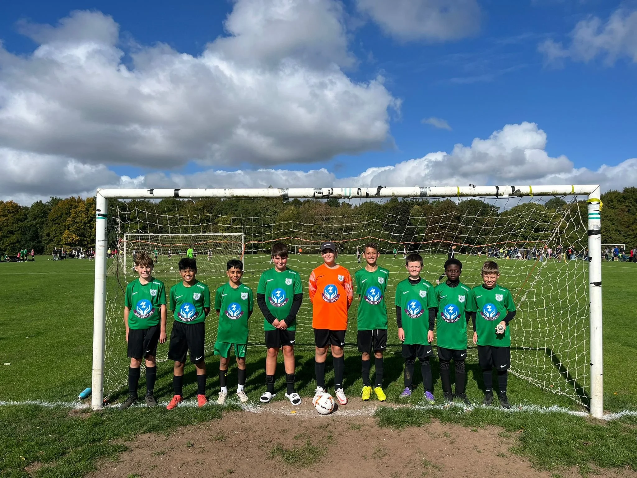 A group of young football players standing in front of a net on a grassy field, with a bright blue sky and clouds overhead.