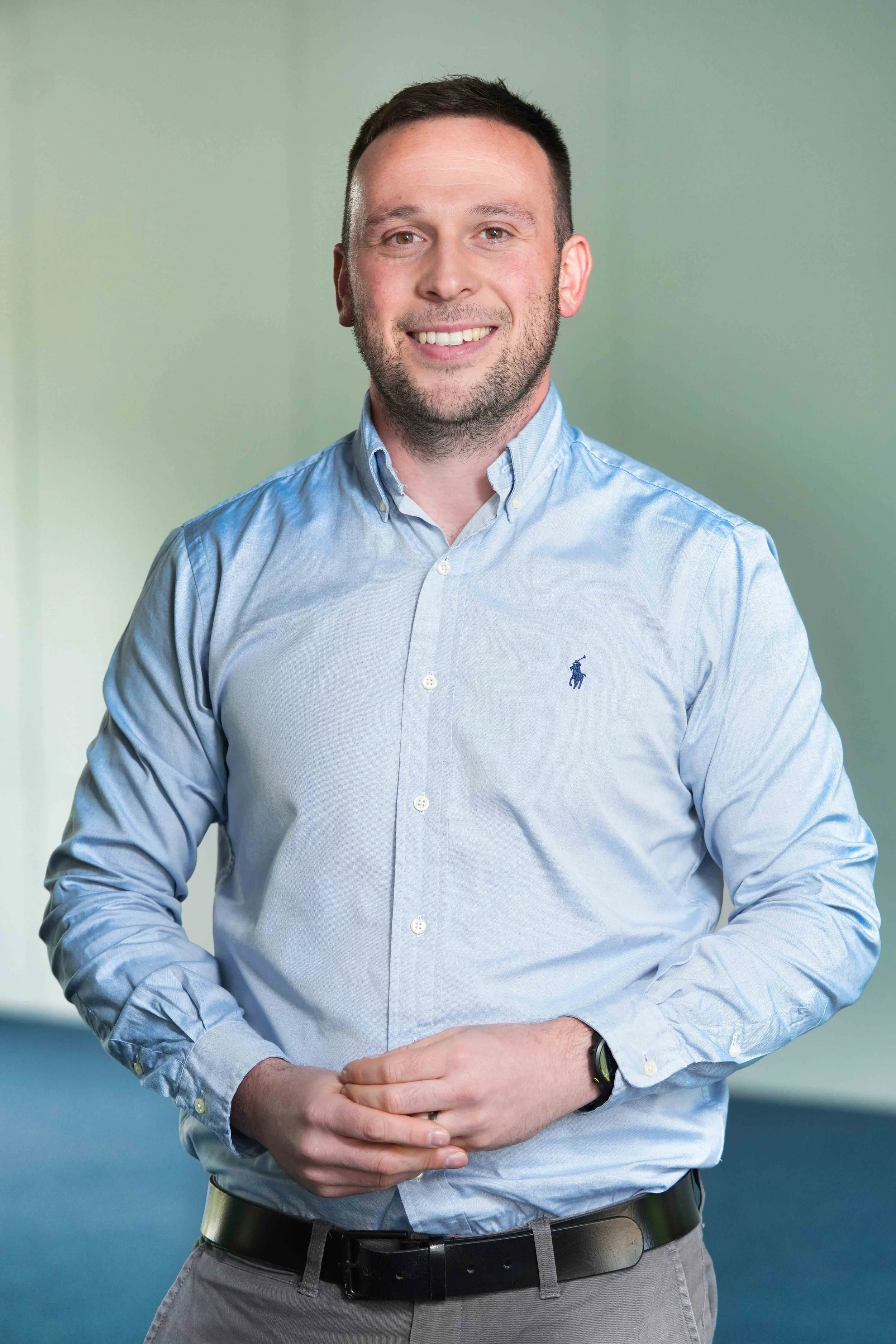 A man wearing a light blue button-up shirt, dark grey pants, and a black watch, standing against a green background with a slight smile.