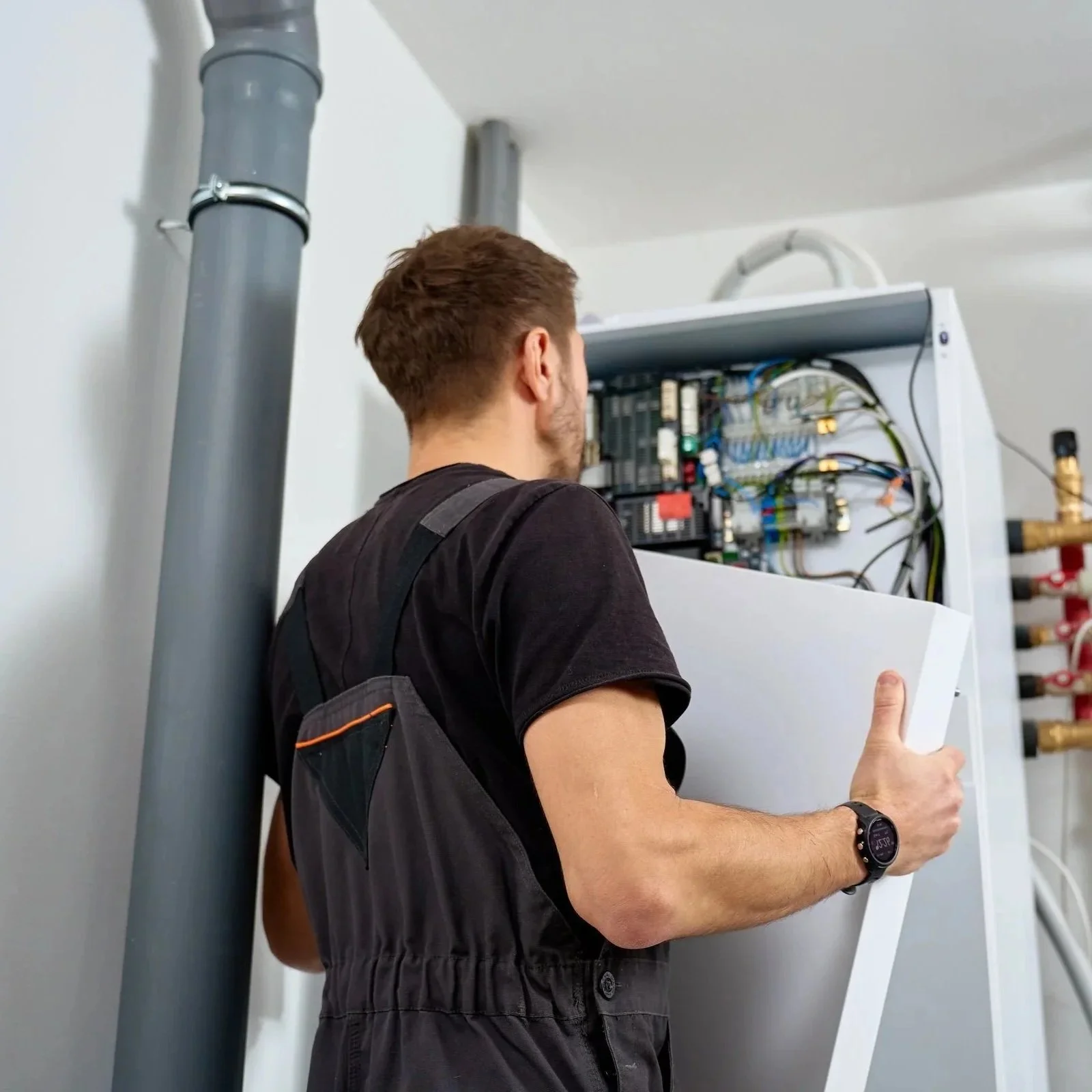 A man wearing a black shirt and a watch is working on an electrical panel or circuit box on a white wall. He is holding a piece of paper or a manual while connected to the panel.