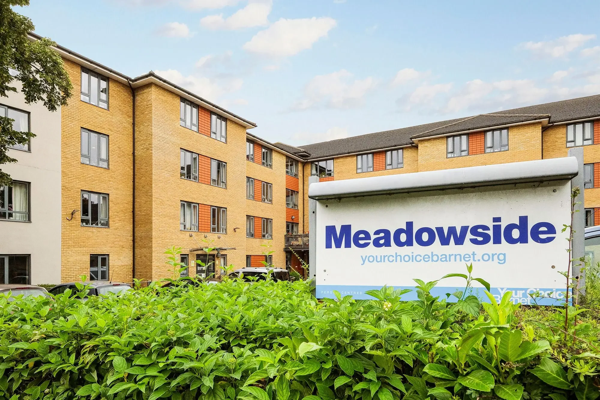 Apartment building with multiple stories and yellow brick exterior, with some red accents, under a partly cloudy sky; foreground shows green plants and a sign that reads 'Meadowside yourchoicebarnet.org'.