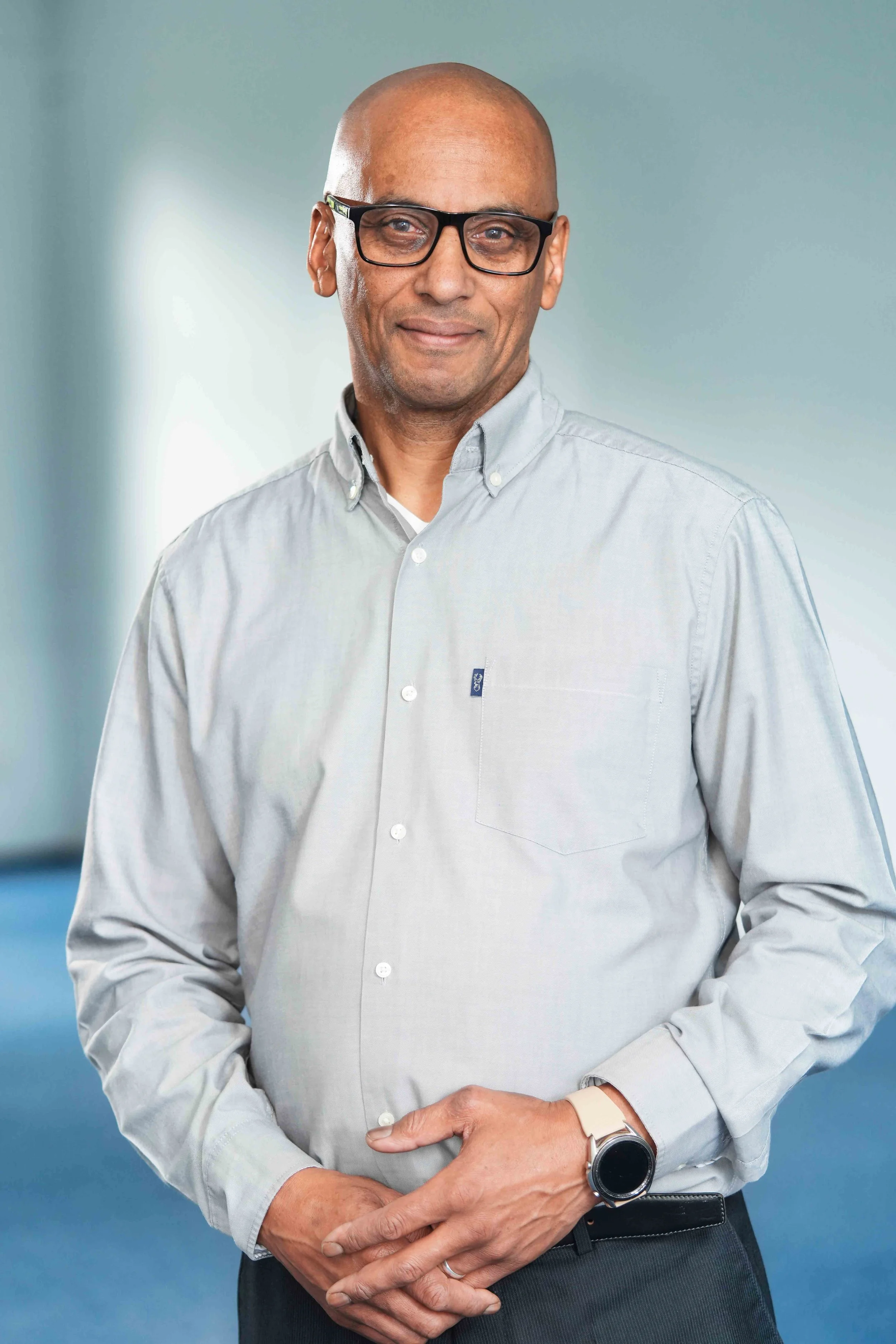 A man with glasses wearing a light grey button-up shirt and a watch, standing against a plain background.