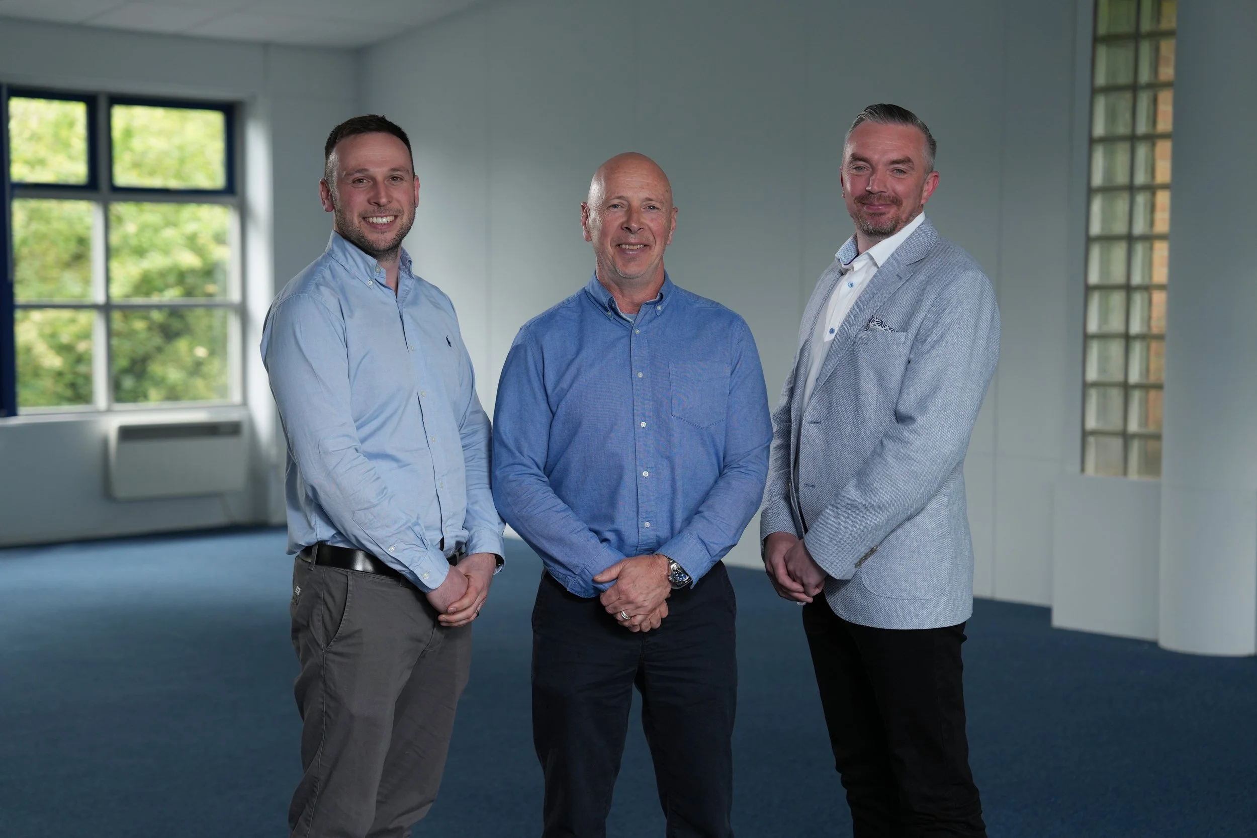 Three men standing together inside a room with large windows showing greenery outside. They are dressed in business casual attire and are smiling at the camera.