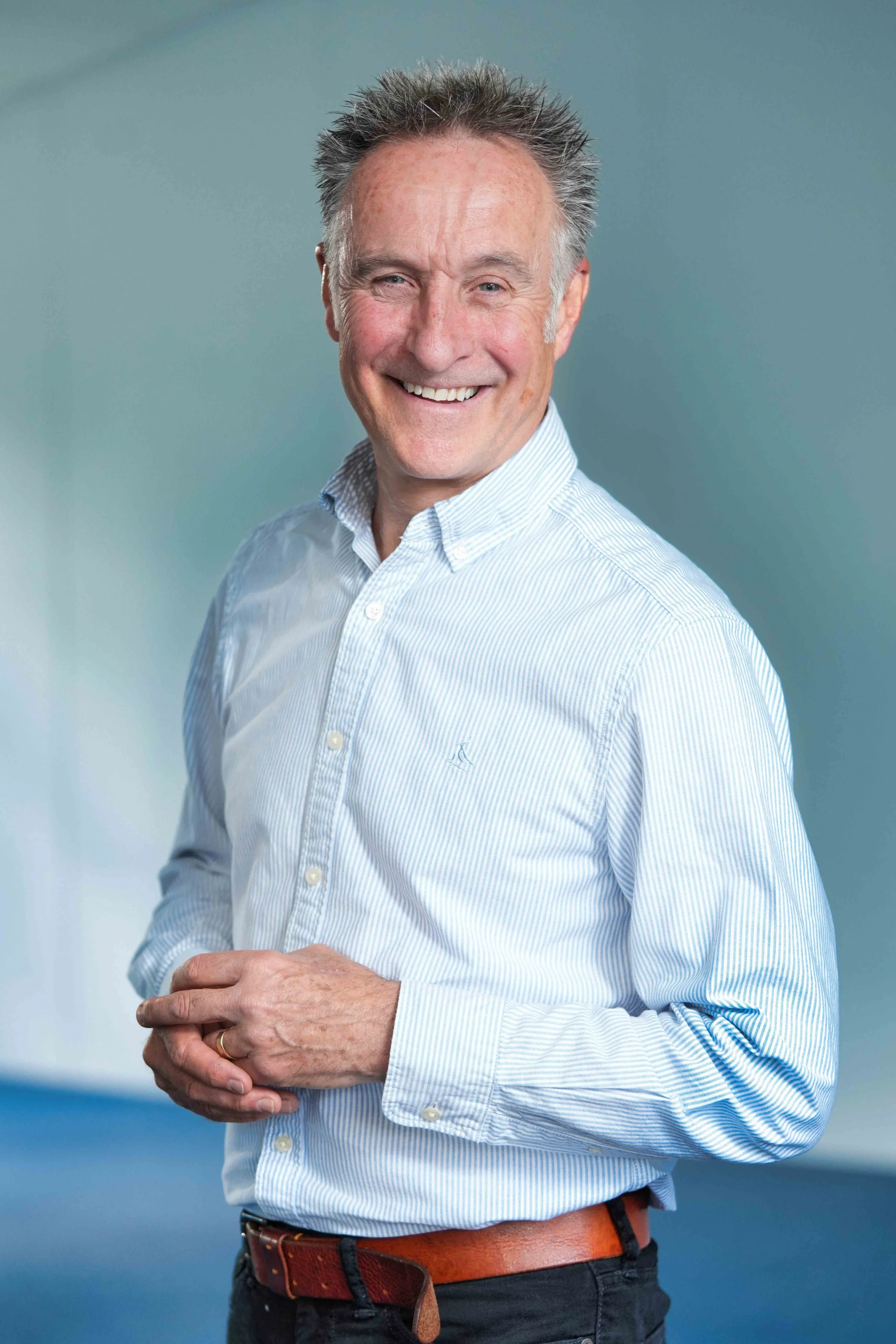 A smiling man wearing a light blue striped button-up shirt, dark pants, and a brown belt, standing against a blue background.