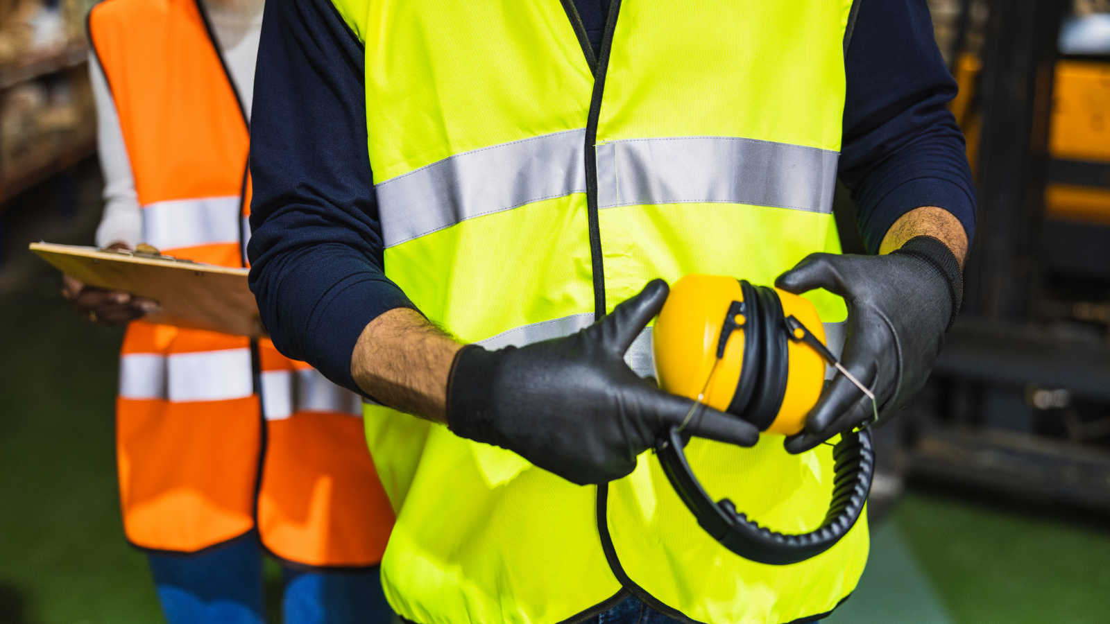 A worker in a neon yellow safety vest and black gloves holding yellow protective earmuffs, with another worker in an orange safety vest holding a clipboard in the background.