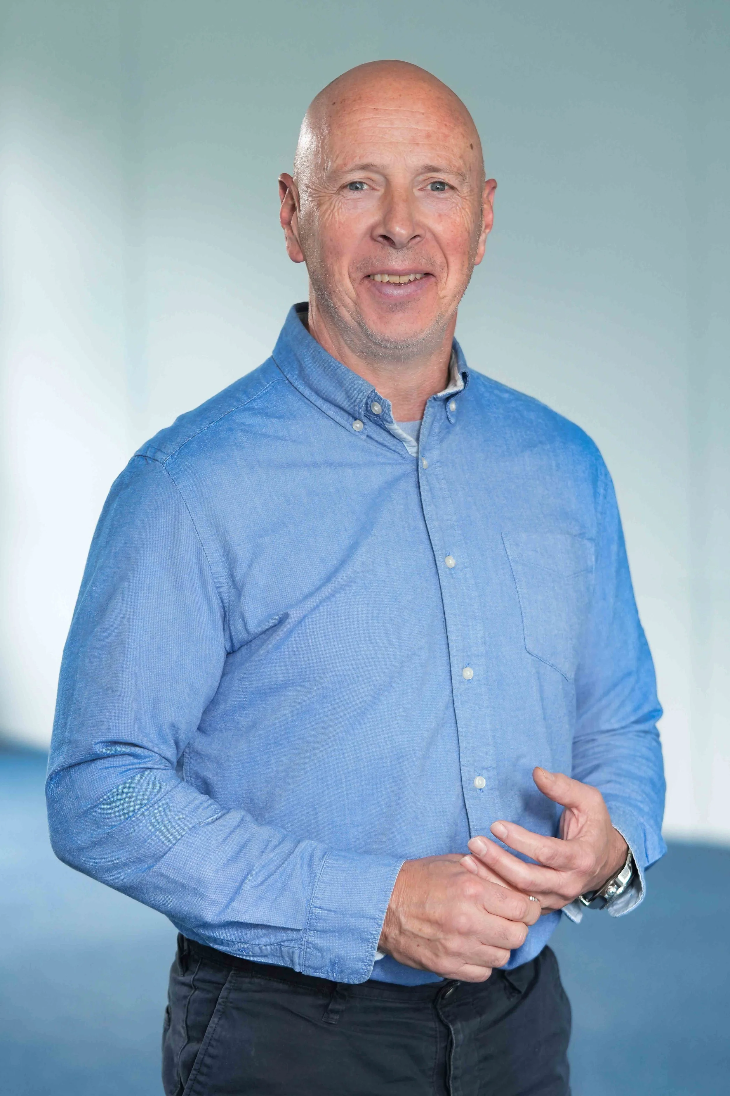 A man wearing a blue button-down shirt, standing against a light blue background.