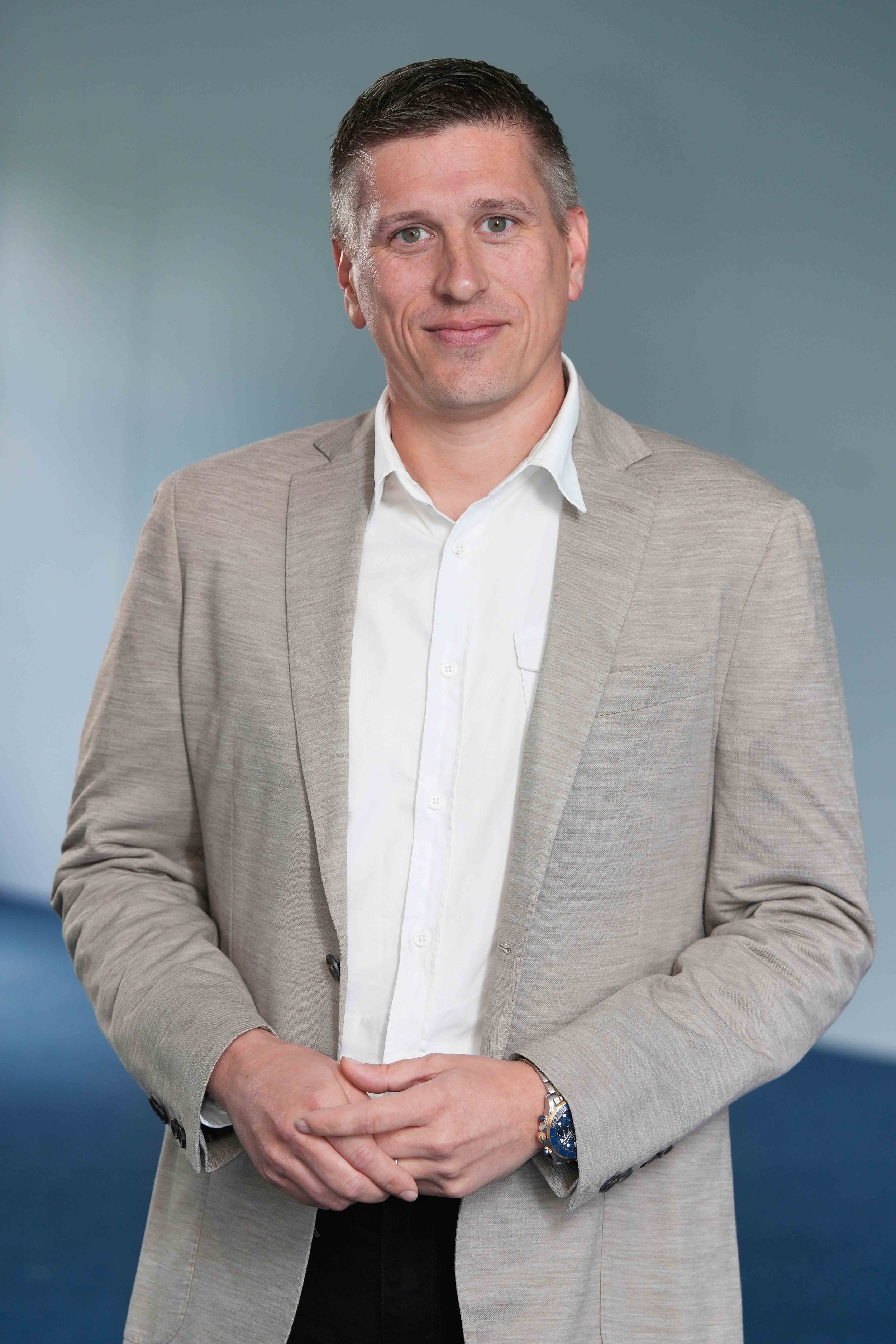 A man in a beige blazer and white shirt standing against a gray background, with clasped hands and a watch on his left wrist.