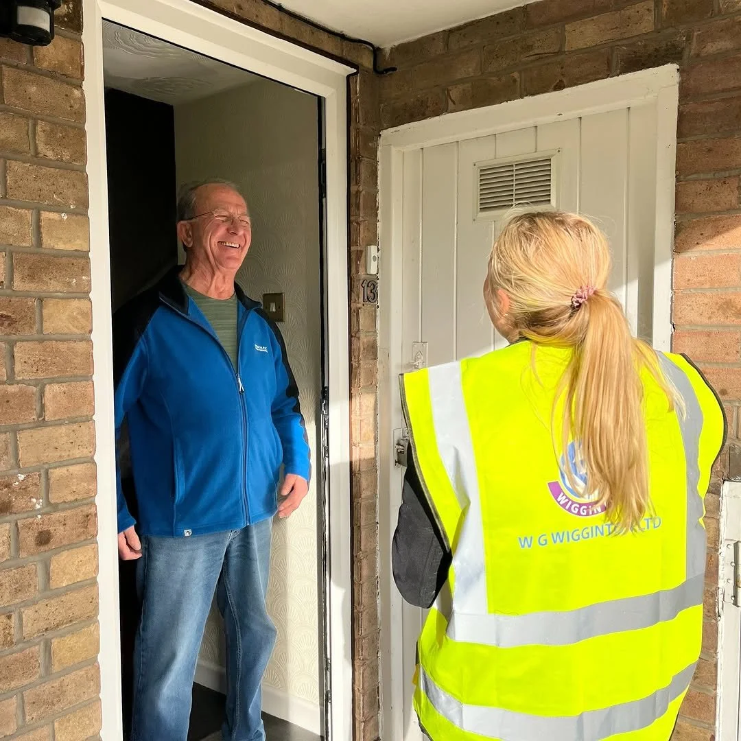 A woman in a bright yellow safety vest speaking with an elderly man standing at his doorway, both smiling.