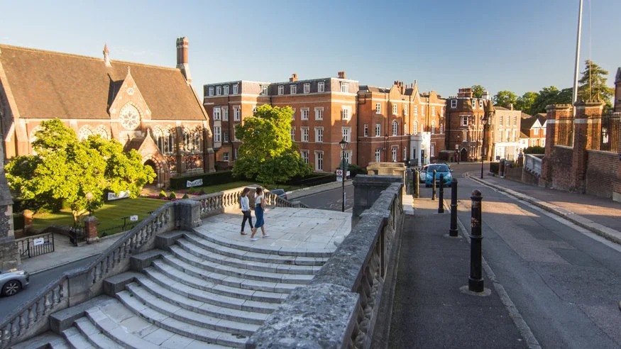 People walking down the steps of a historic stone staircase in a city with red brick buildings and a church with a large stained glass window, green trees, and a sunny sky.
