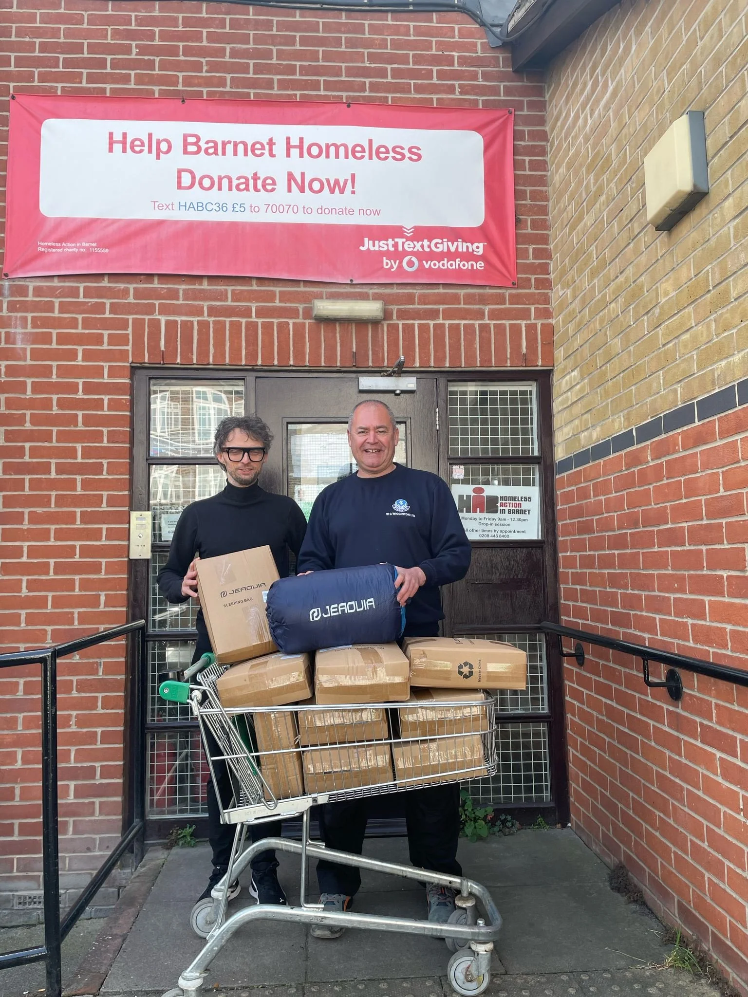 Two men standing in front of a door holding a shopping cart filled with cardboard boxes. One man is wearing glasses and a black turtleneck, the other man is smiling and wearing a black sweatshirt. There is a pink banner above them with the text 'Help Barnet Homeless Donate Now!' and information about donations. The brick building has a door with a small window and signs about homelessness support.