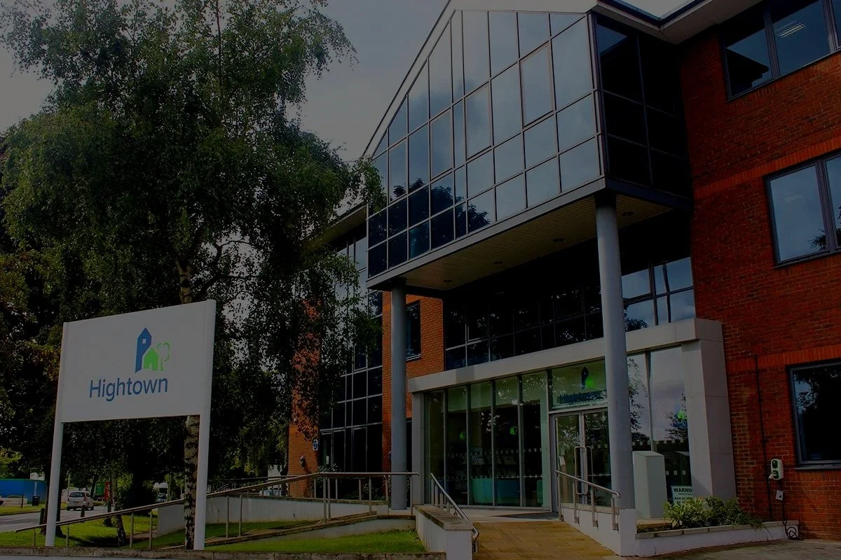 Exterior of a modern office building with glass windows and a sign that reads "Hightown" with a logo of a blue and green house. Trees are visible in the background.