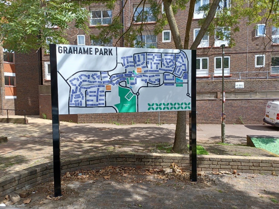 Map of Grahame Park showing streets, pathways, green spaces, and recreational areas, set outdoors with trees and an urban building in the background.