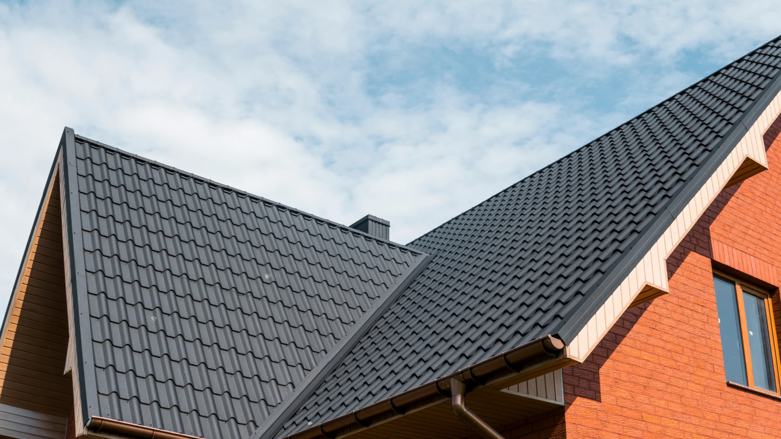 Close-up of a house roof with gray metal tiles, brick exterior wall, a window, and a cloudy sky.
