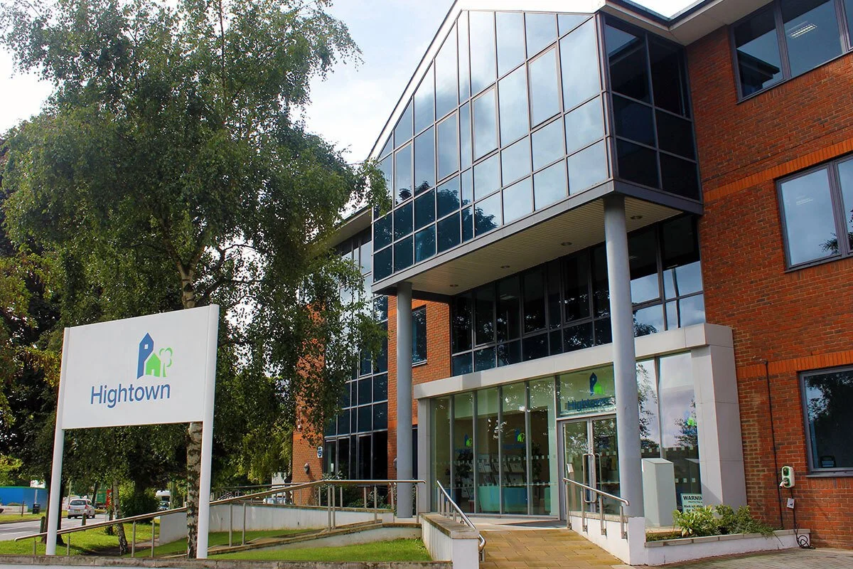 Exterior of a modern brick building with large glass windows and an entrance ramp. There's a sign that reads 'Hightown' with a logo of a house and a tree.