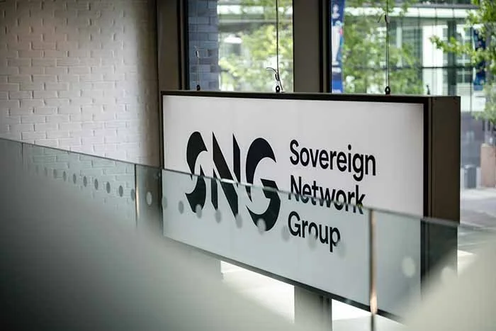Sign for the Sovereign Network Group (SNG) hanging inside a building with large windows and a view of trees outside.