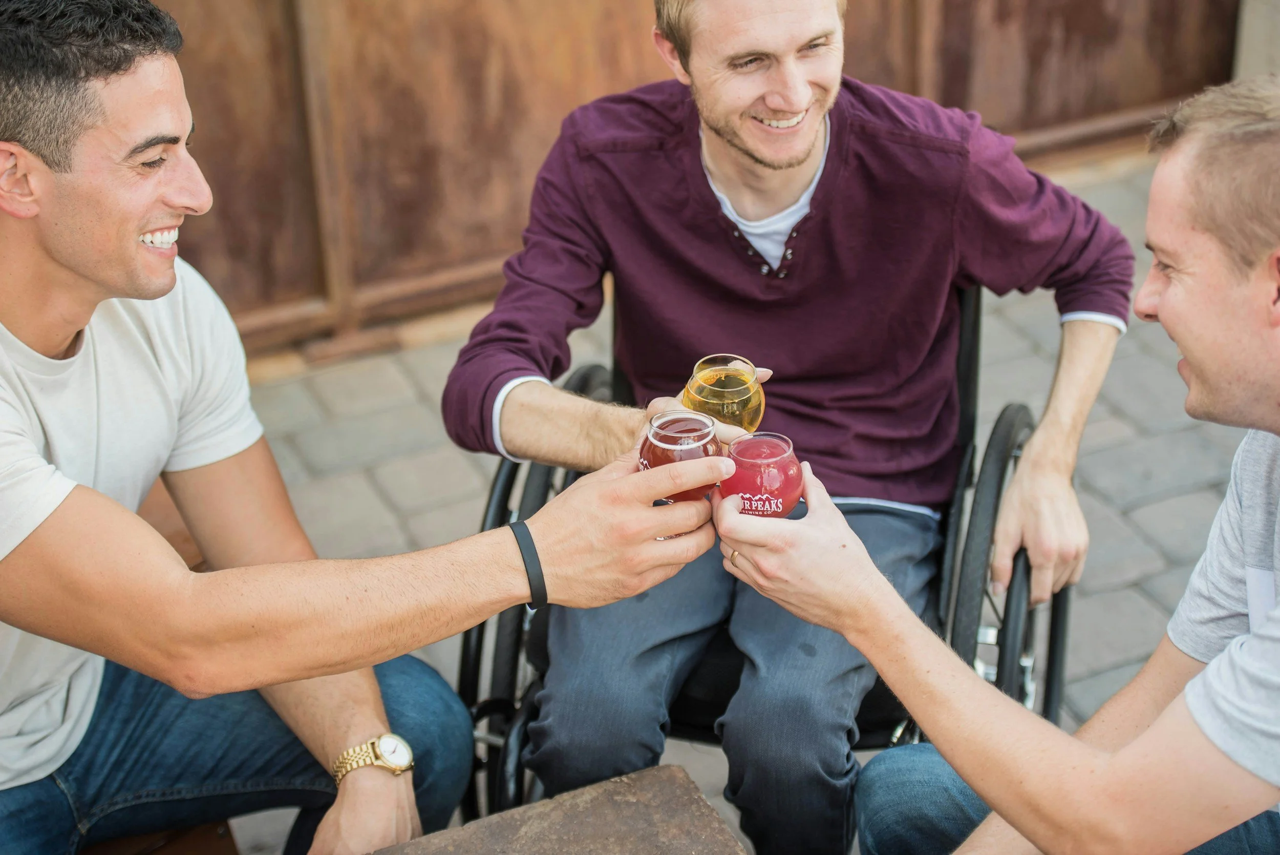 Four men clinking drinks together at an outdoor cafe, one of whom is in a wheelchair, all smiling and enjoying each other's company.