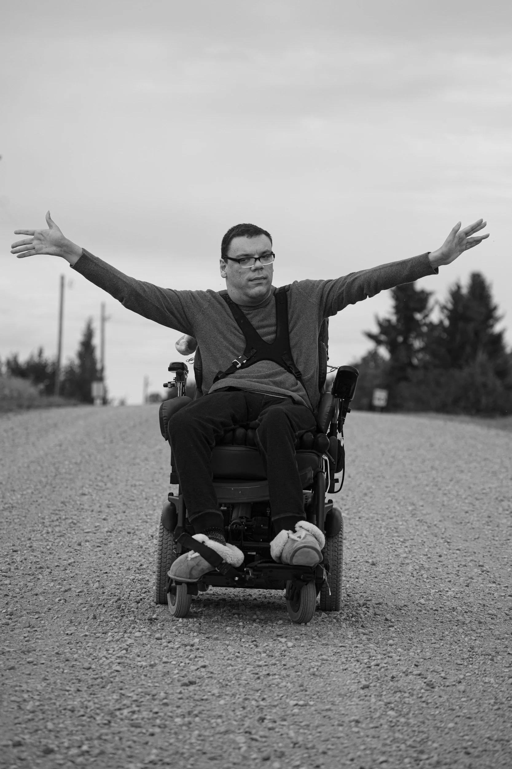Man in a wheelchair with arms outstretched on a gravel road, with trees and utility poles in the background, black and white photo.