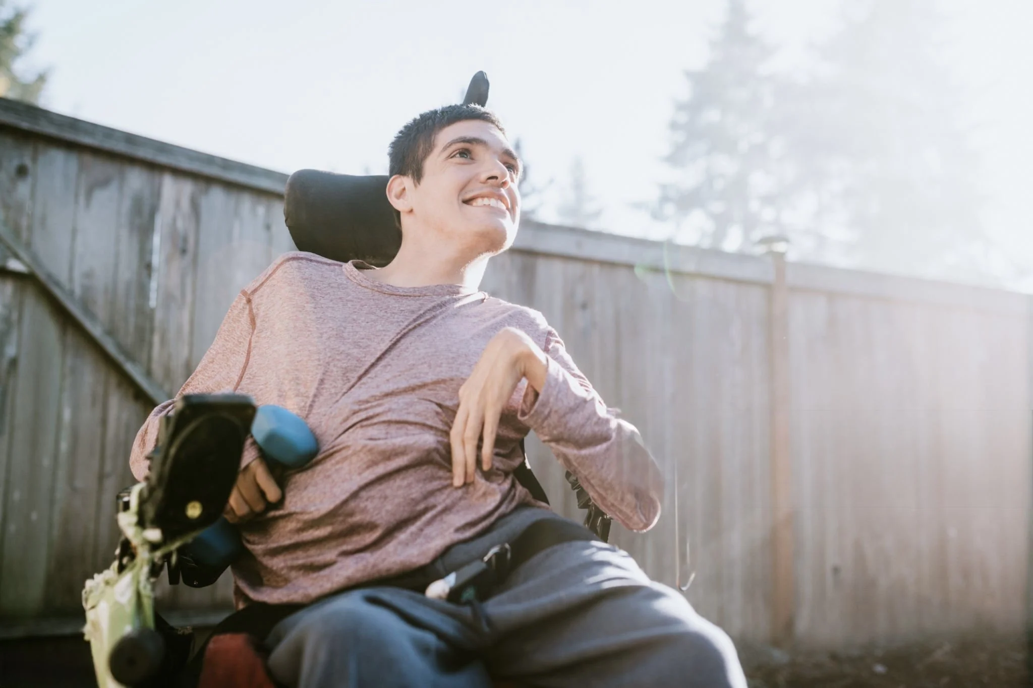 Young man in a wheelchair outdoors, smiling, with sunlight shining through trees in the background.