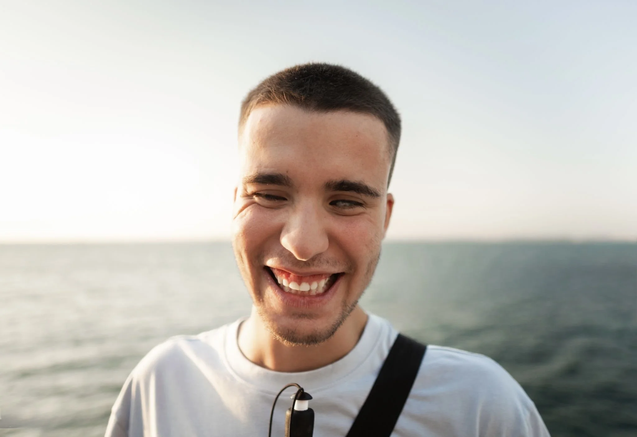 Young man with short hair smiling outdoors near water, wearing a white shirt and carrying a black strap.