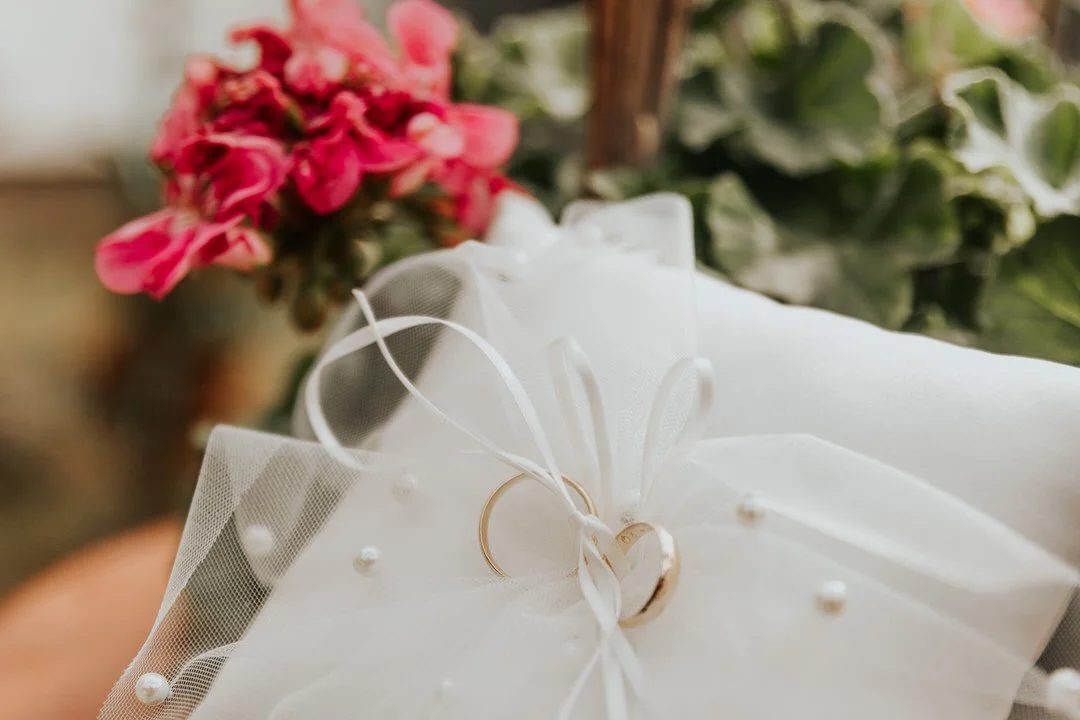 Close-up of a wedding ring resting on white tulle with pearl accents and pink flowers in the background.