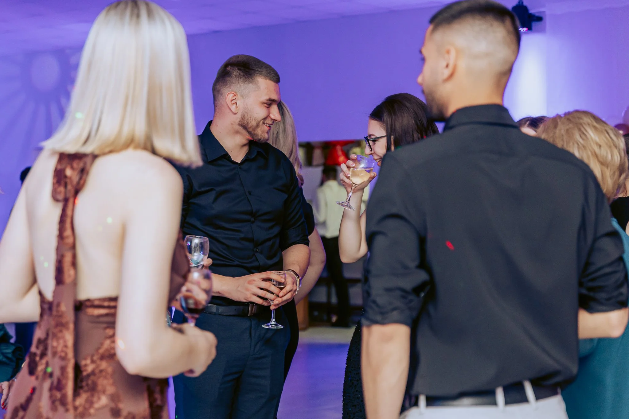 Group of people socializing at a party with dim, colorful lighting. Some are holding glasses of champagne or wine and engaging in conversations.