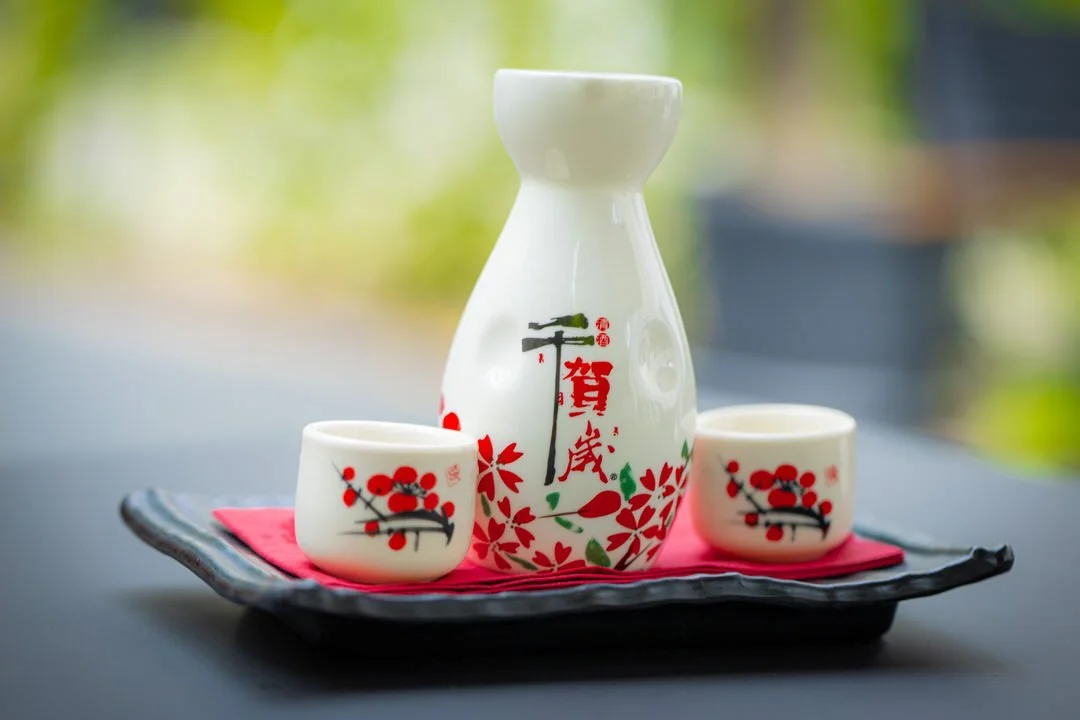 A white sake set with a decanter and two cups, decorated with red and black floral patterns, placed on a black tray with a red cloth underneath.